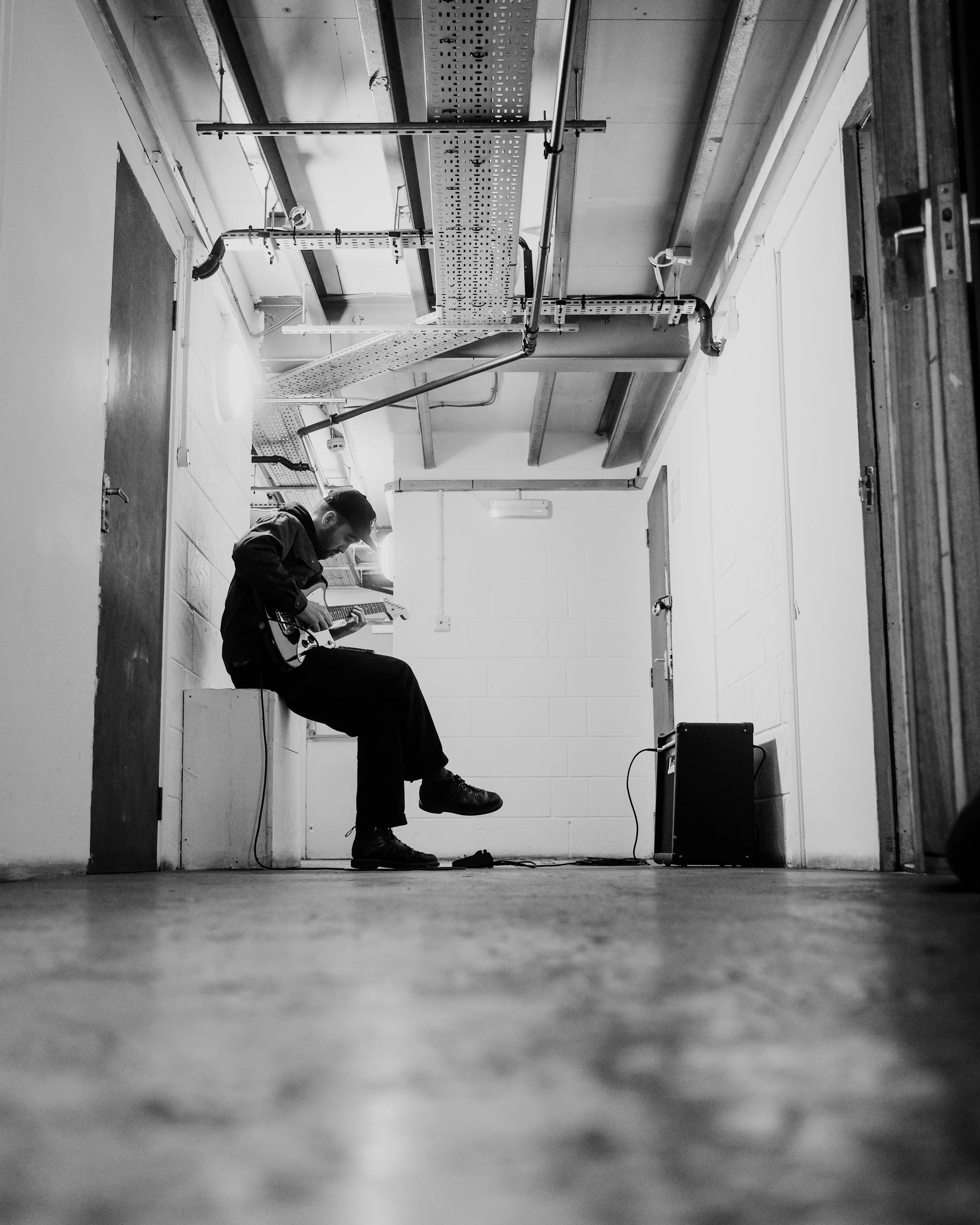 A man sitting on a box in a room, playing an electric guitar. The room has pipes and panels on the ceiling, with a small amplifier on the floor nearby.