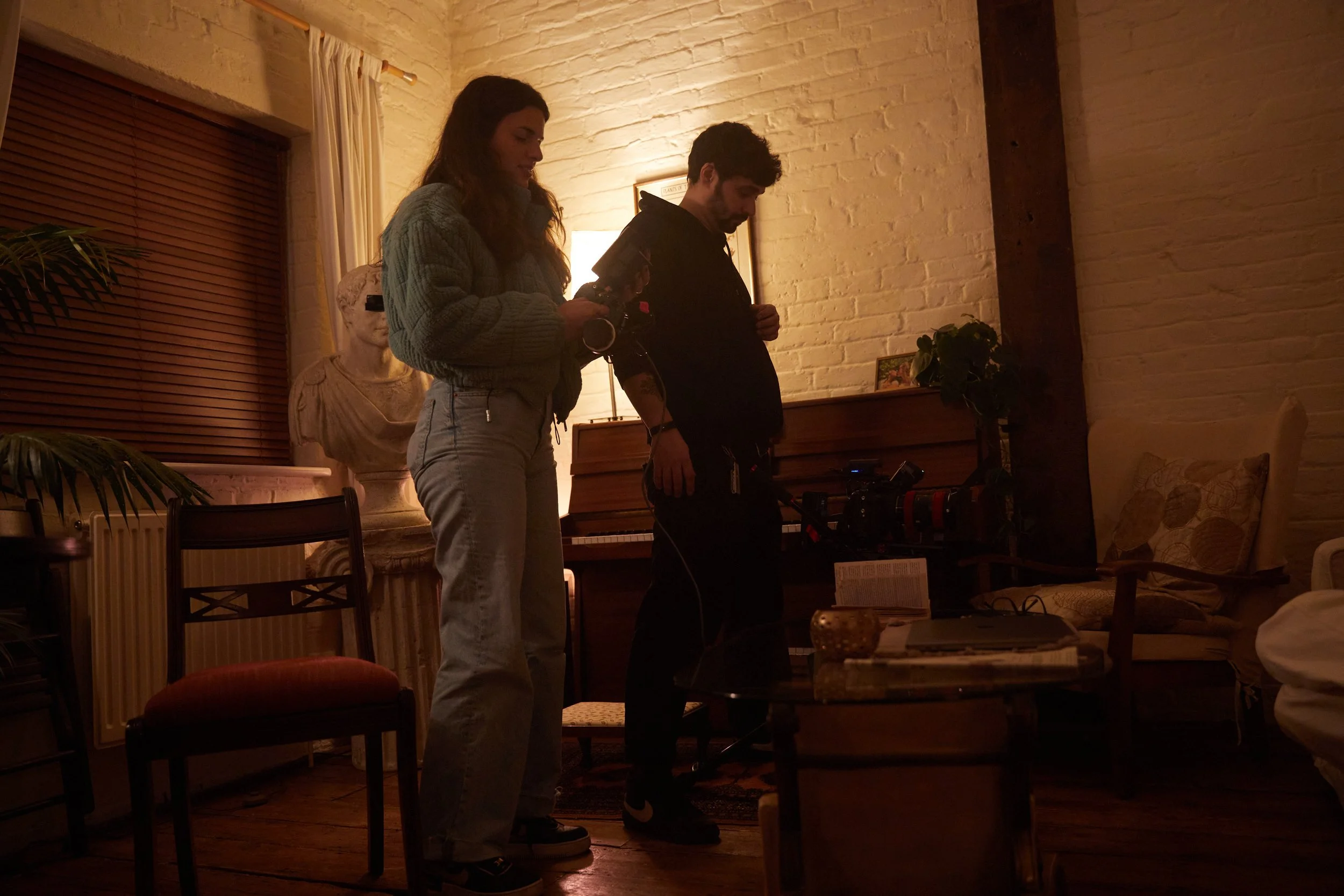 Three people working on a film or photography set in a warmly lit living room with a brick wall, a piano, and various furniture and decorations.