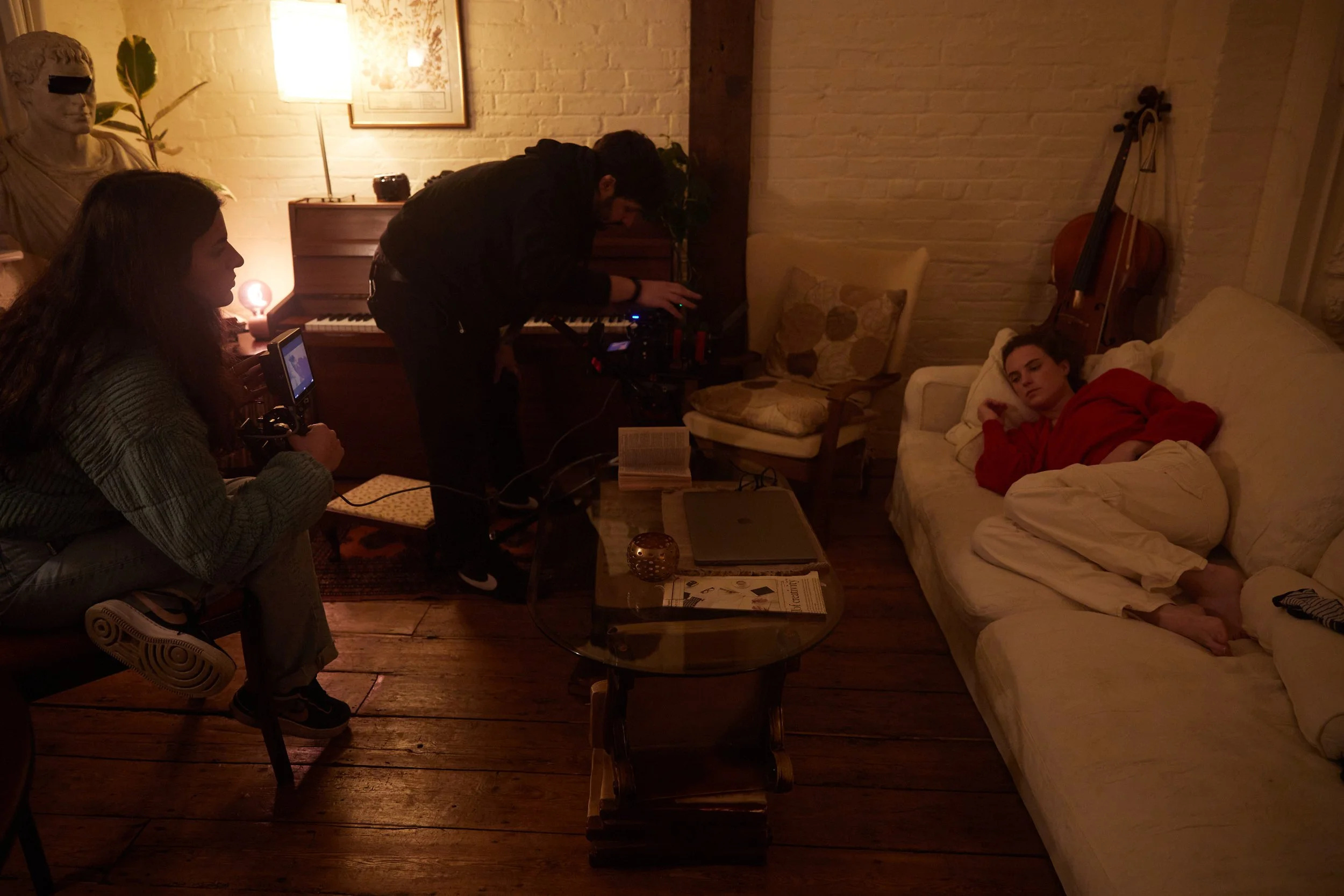 Film crew preparing a scene in a cozy living room. A young man in a red shirt lies on a cream-colored couch, looking at the camera. A woman holds a camera, and a man arranges equipment near a piano and a sitting area.