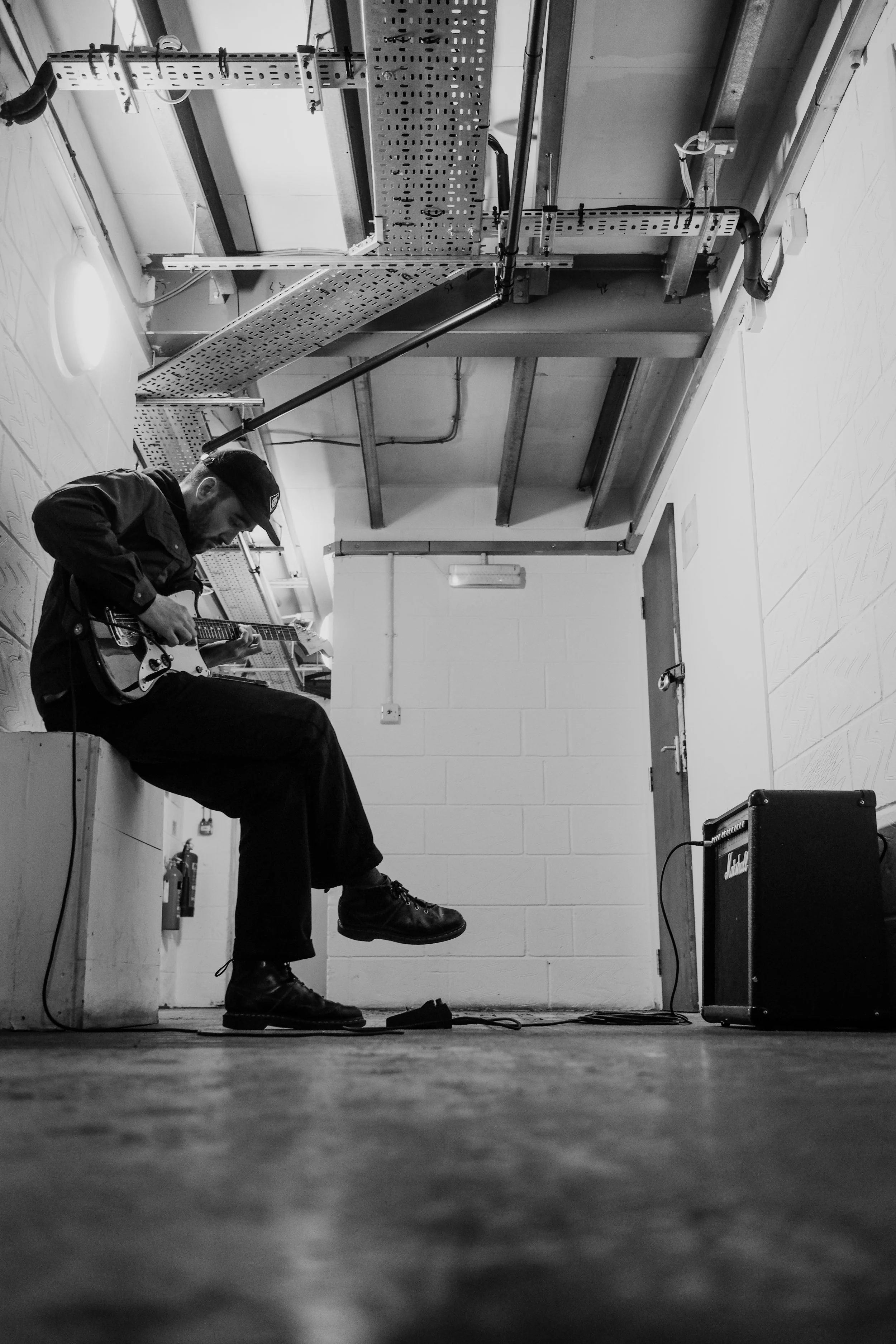 A man sitting on a box playing an electric guitar in a backstage or rehearsal room, with a guitar amplifier nearby, in black and white.