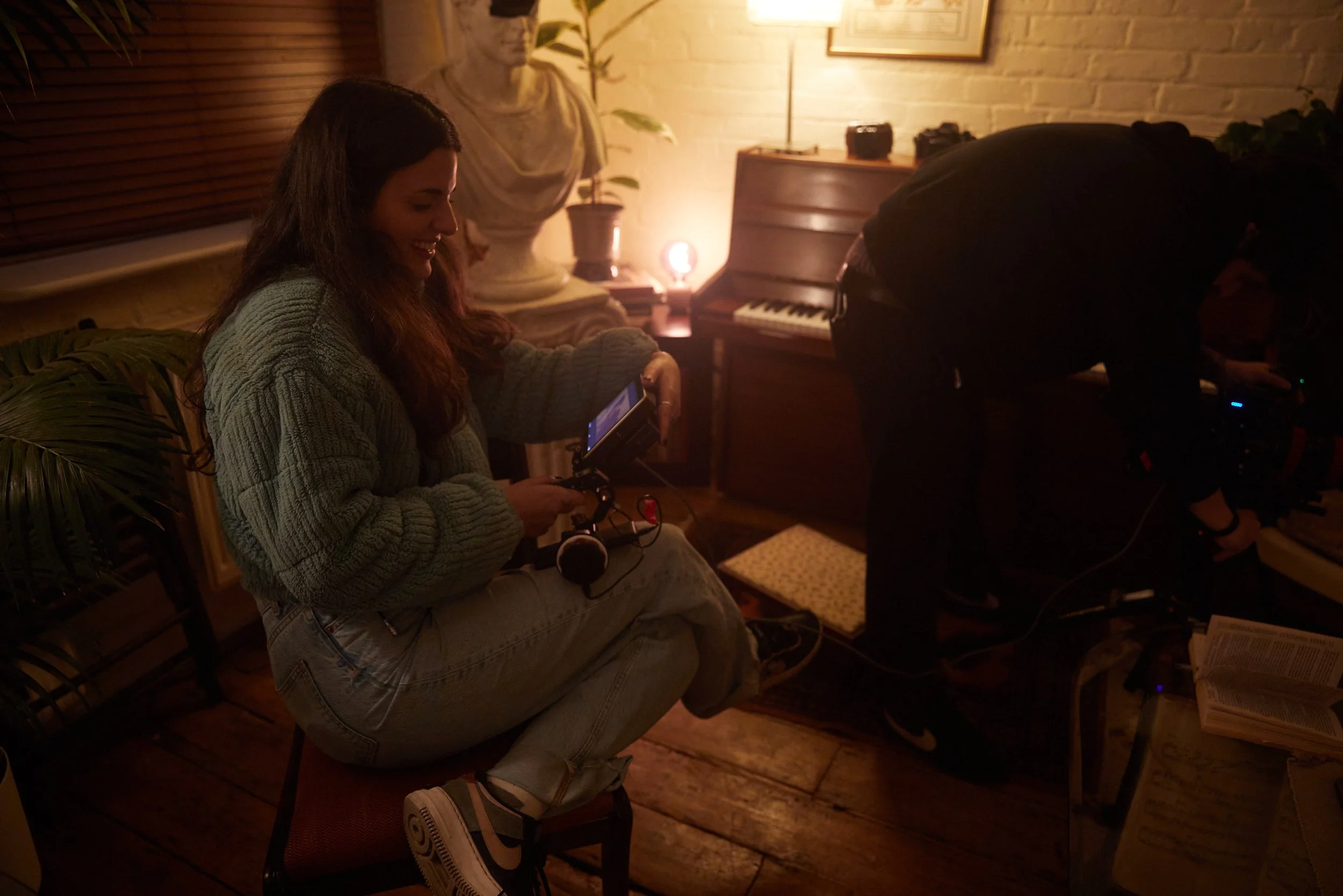 A woman seated on a chair in a dimly lit room, holding a camera monitor, with a man adjusting camera equipment nearby, and a classical statue, piano, and framed picture on the wall in the background.