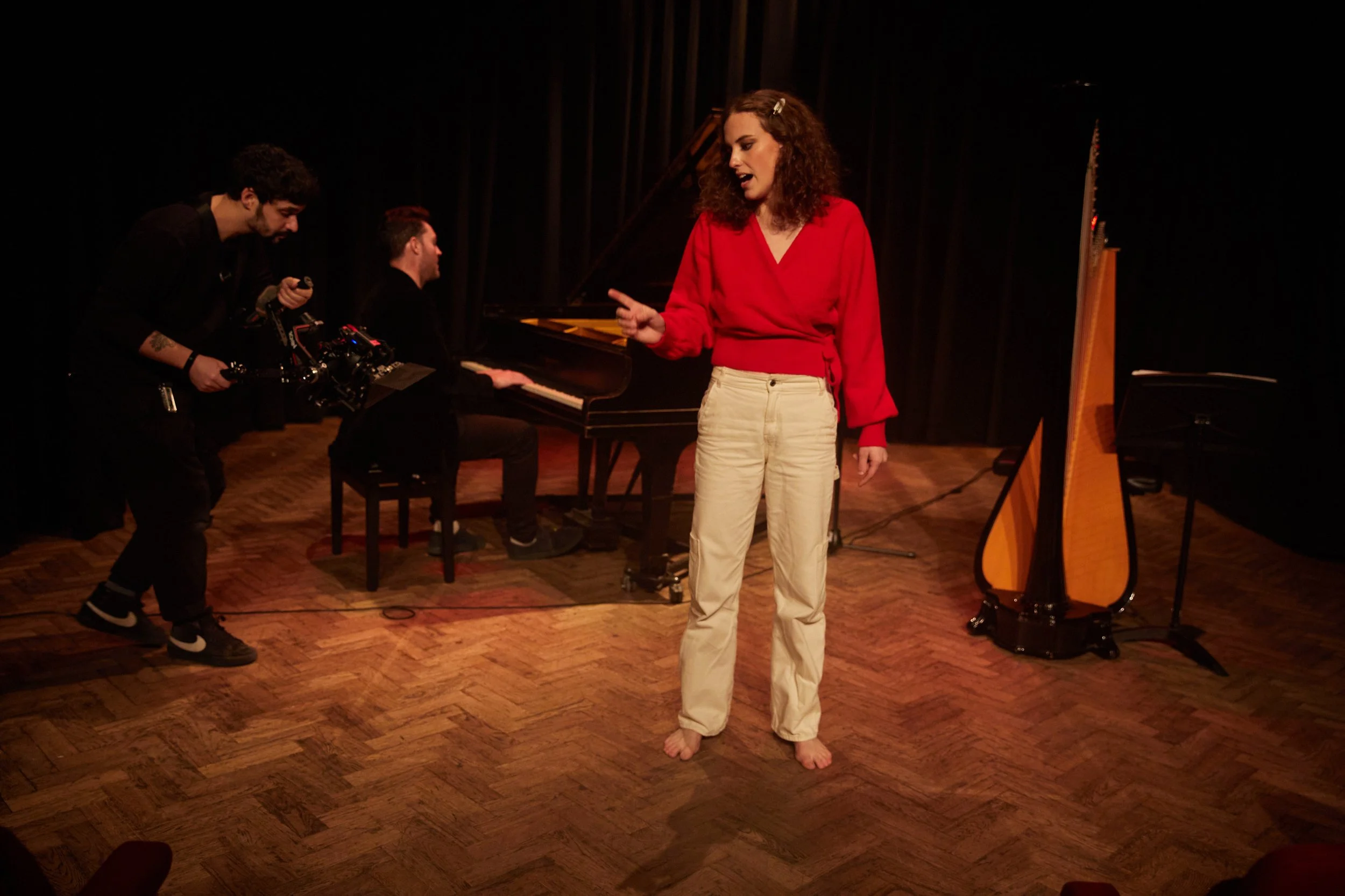 A woman in a red top and beige pants performing on stage, with a pianist and a camera crew filming her, a grand piano and harp in the background.