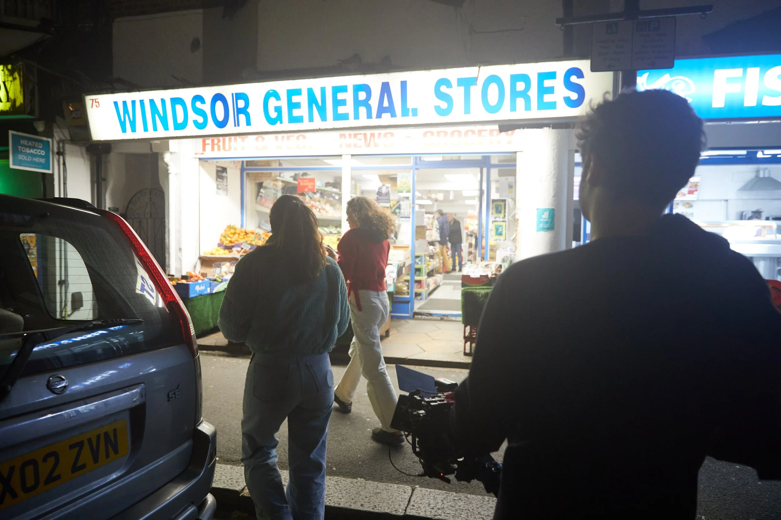 Nighttime scene outside Windsor General Stores with customers and a camera person in the foreground, store front displaying fresh produce, and bright signage.