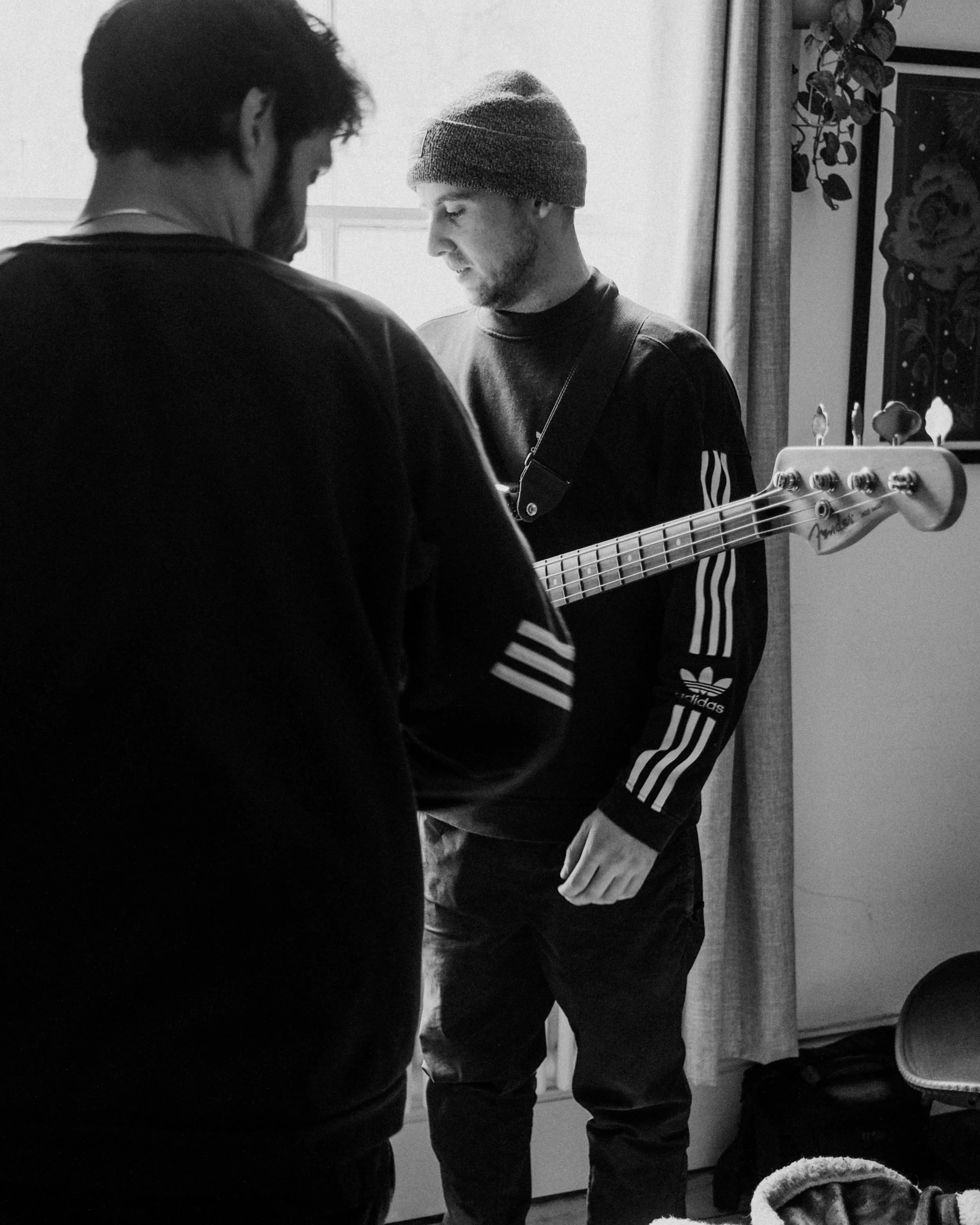 Two men, one with a guitar, indoors near a window with curtains.