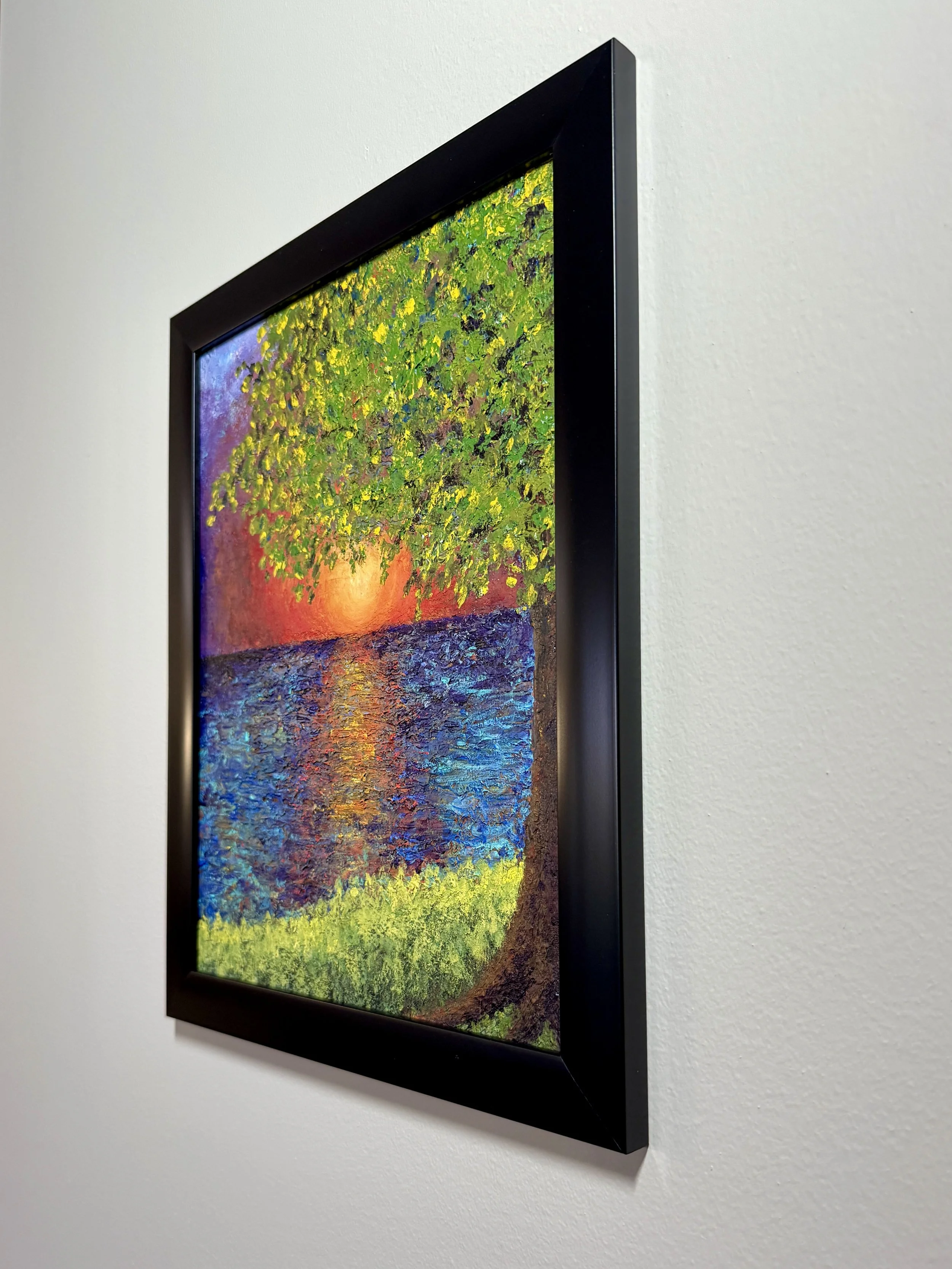 A painting of a vibrant tree with green leaves reflecting sunlight on a water body, displayed on a white wall with a black frame.