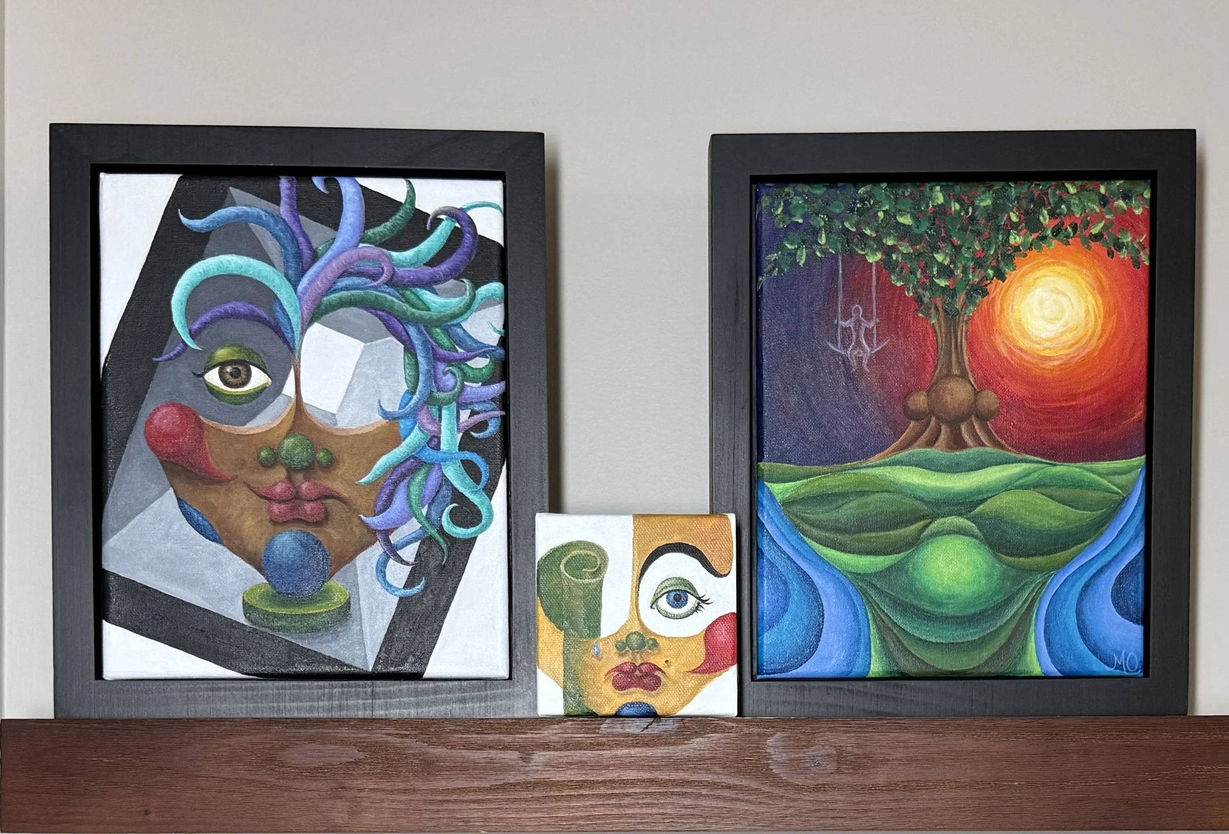 A display of four colorful art pieces on a wall, including paintings of a face with abstract hair, a tree with a sun, a face with a yellow background, and an abstract face with a single eye.