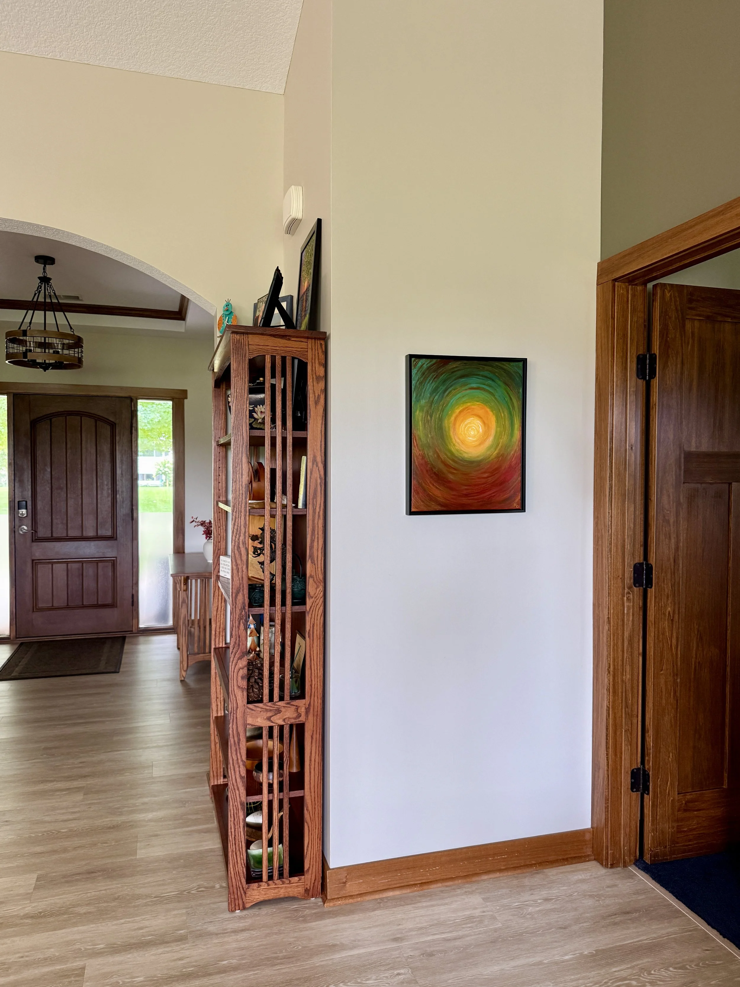 Interior view of a living space with a wooden door, a decorative wooden shelf, colorful art on the wall, and a hallway with a door leading outside, all featuring warm wood accents and hardwood flooring.