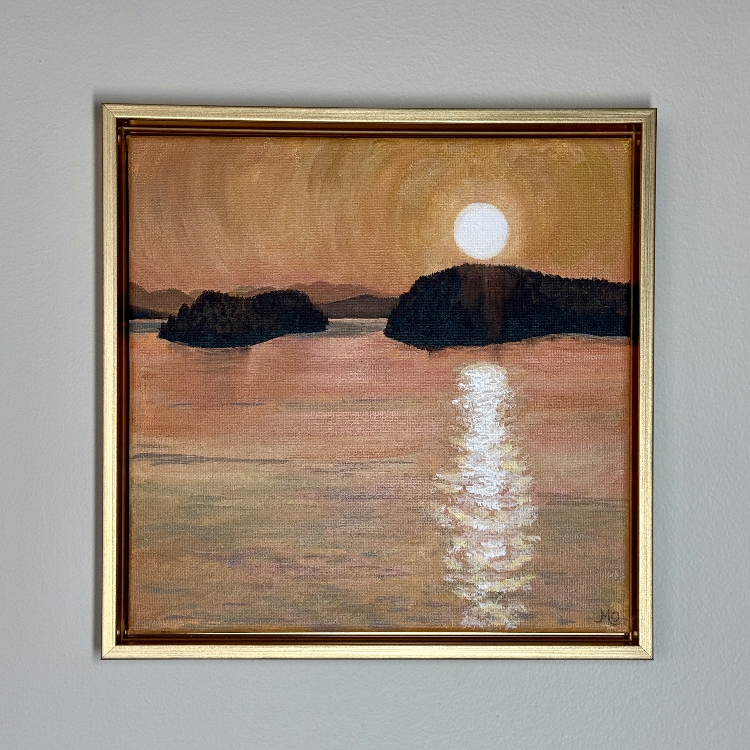 A framed painting of a sunset with reflection on the water, trees in the foreground, and mountains in the background on a gray wall.