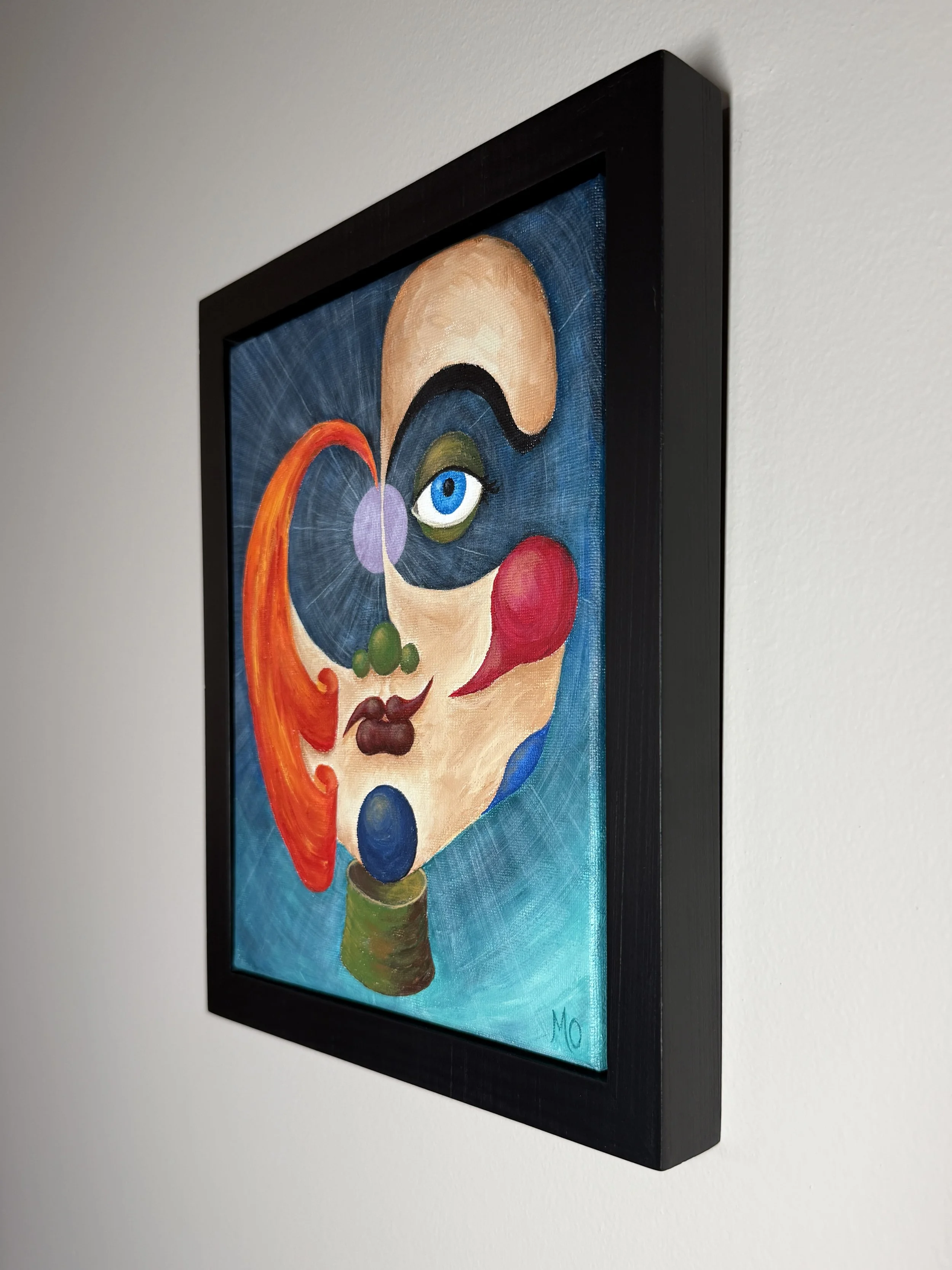 A colorful abstract painting of a woman's face with an eye, red lipstick, and various vivid shapes and colors, framed in black and mounted on a white wall.