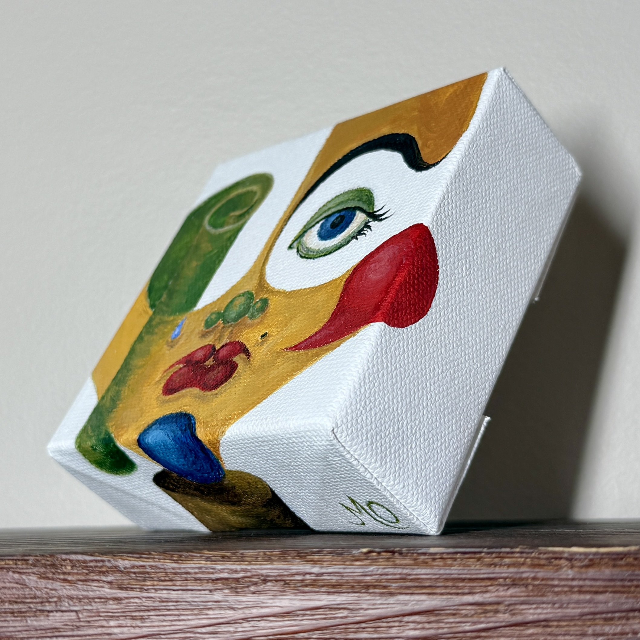 Close-up of a decorated canvas with the face of a clown or jester, featuring makeup and colorful elements, placed on a wooden surface.