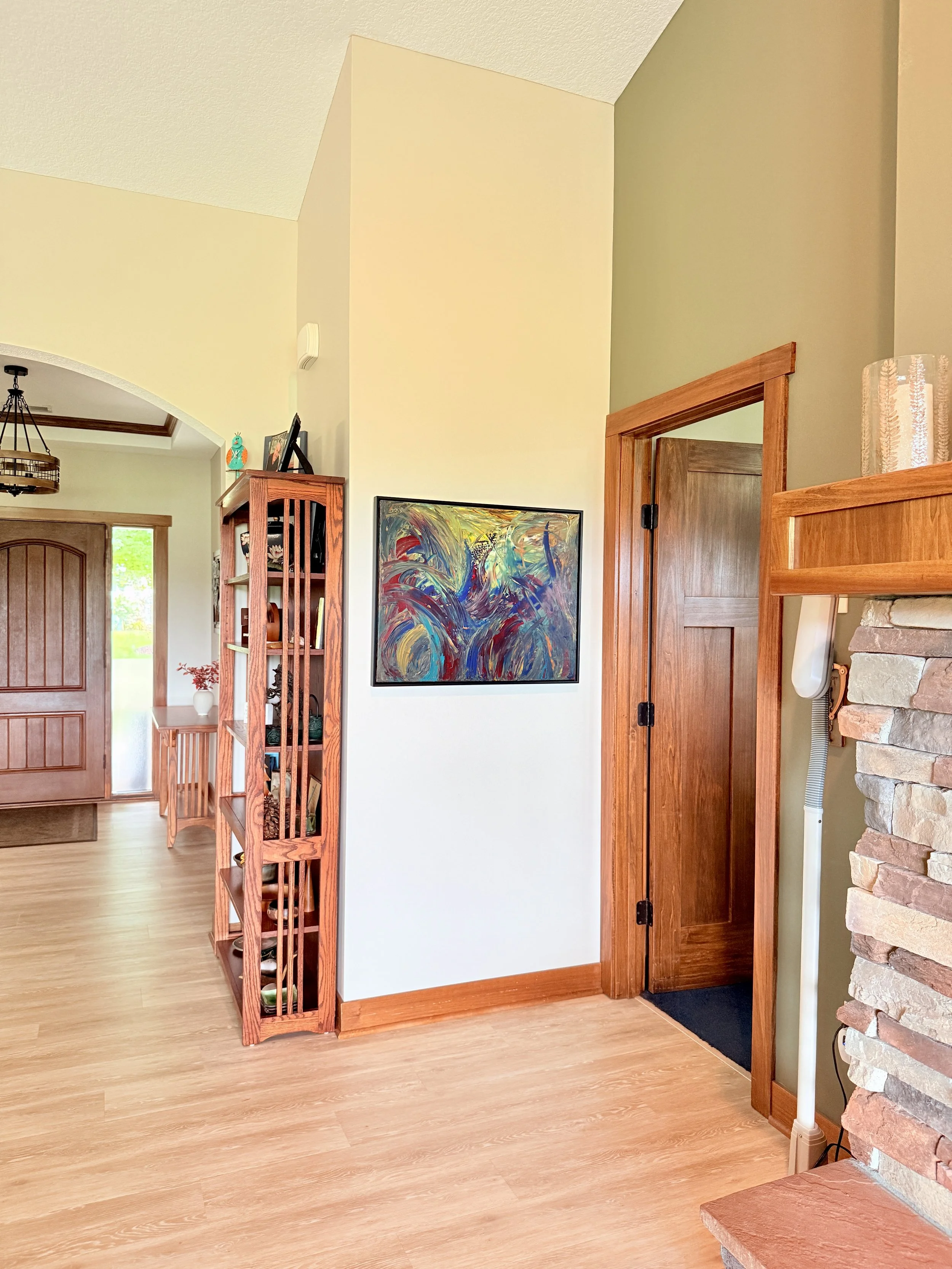 Interior view of a home entryway with wooden floors, a wooden door, a stone fireplace, artwork on the wall, a wooden bookshelf, and decorated shelves with objects.