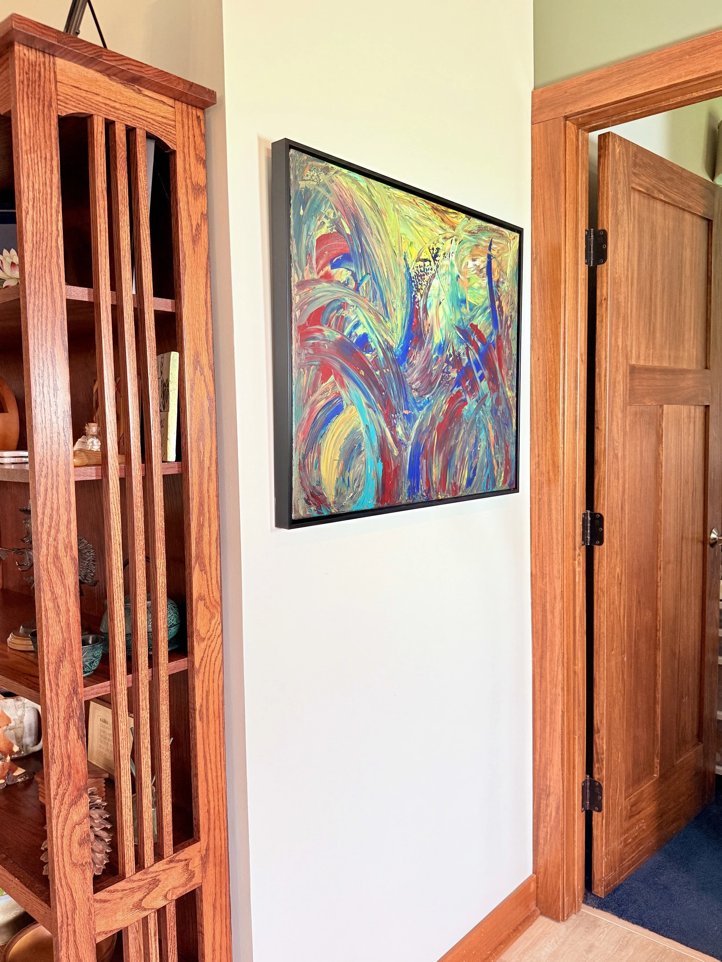 A colorful abstract painting hanging on a white wall next to a wooden door frame with a partially open door. To the left, there is a tall wooden bookshelf with various items.