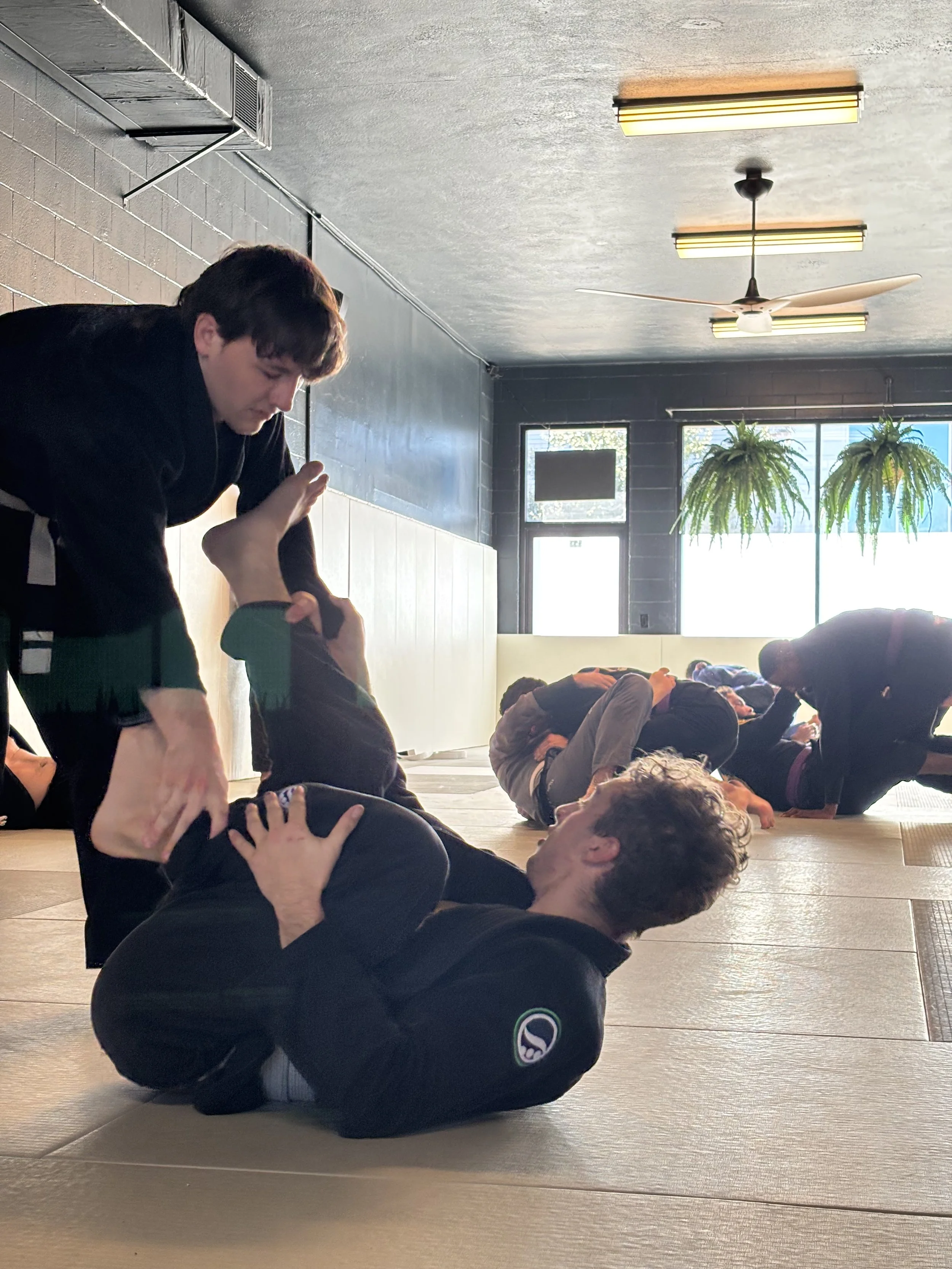 People practicing Jiu-Jitsu on mats in a martial arts gym, with some lying on the floor and others assisting or observing.