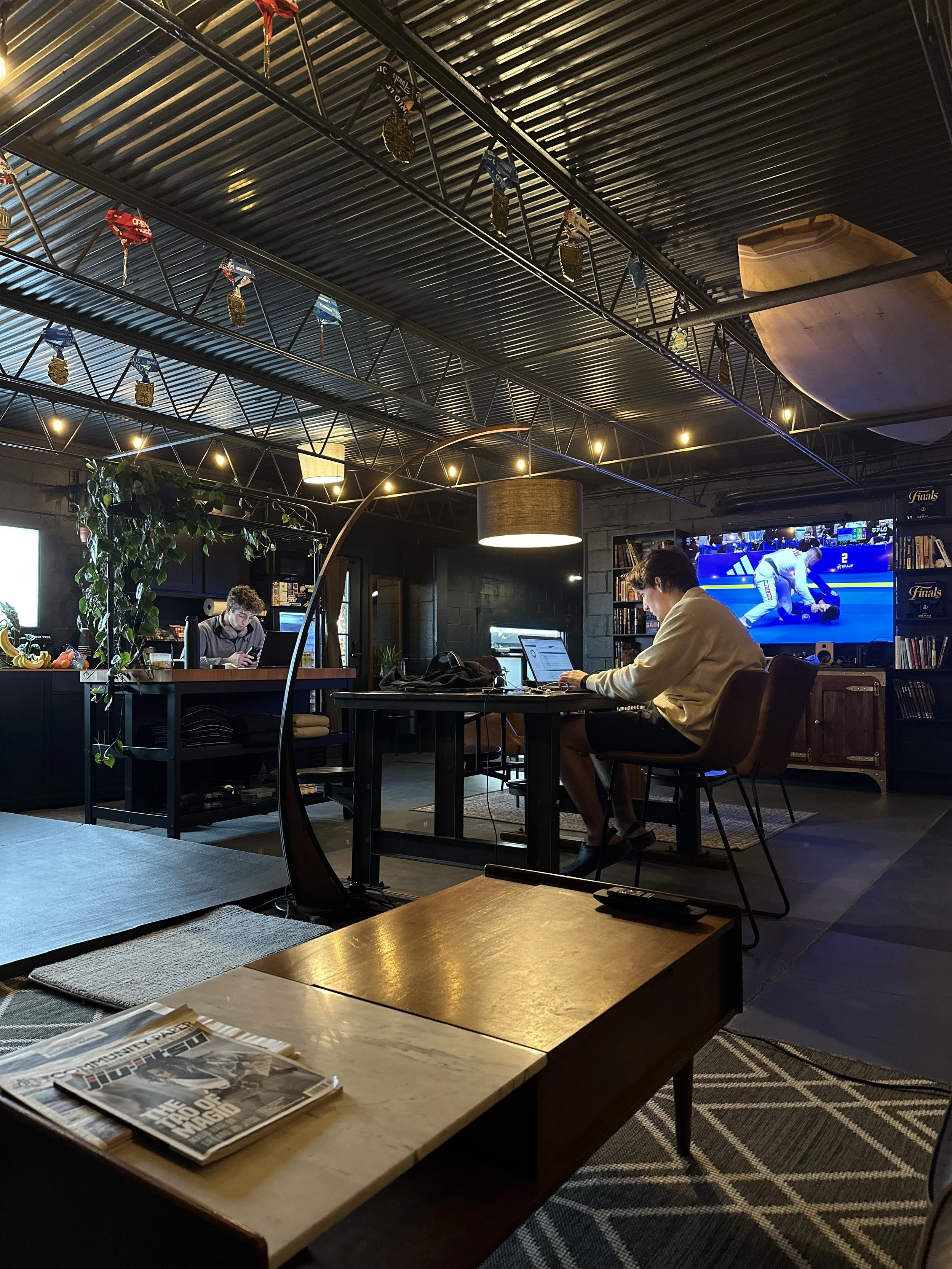 People working on laptops in a modern, cozy indoor space with a dark ceiling, bookshelves, and a large TV showing a jiu jitsu match.