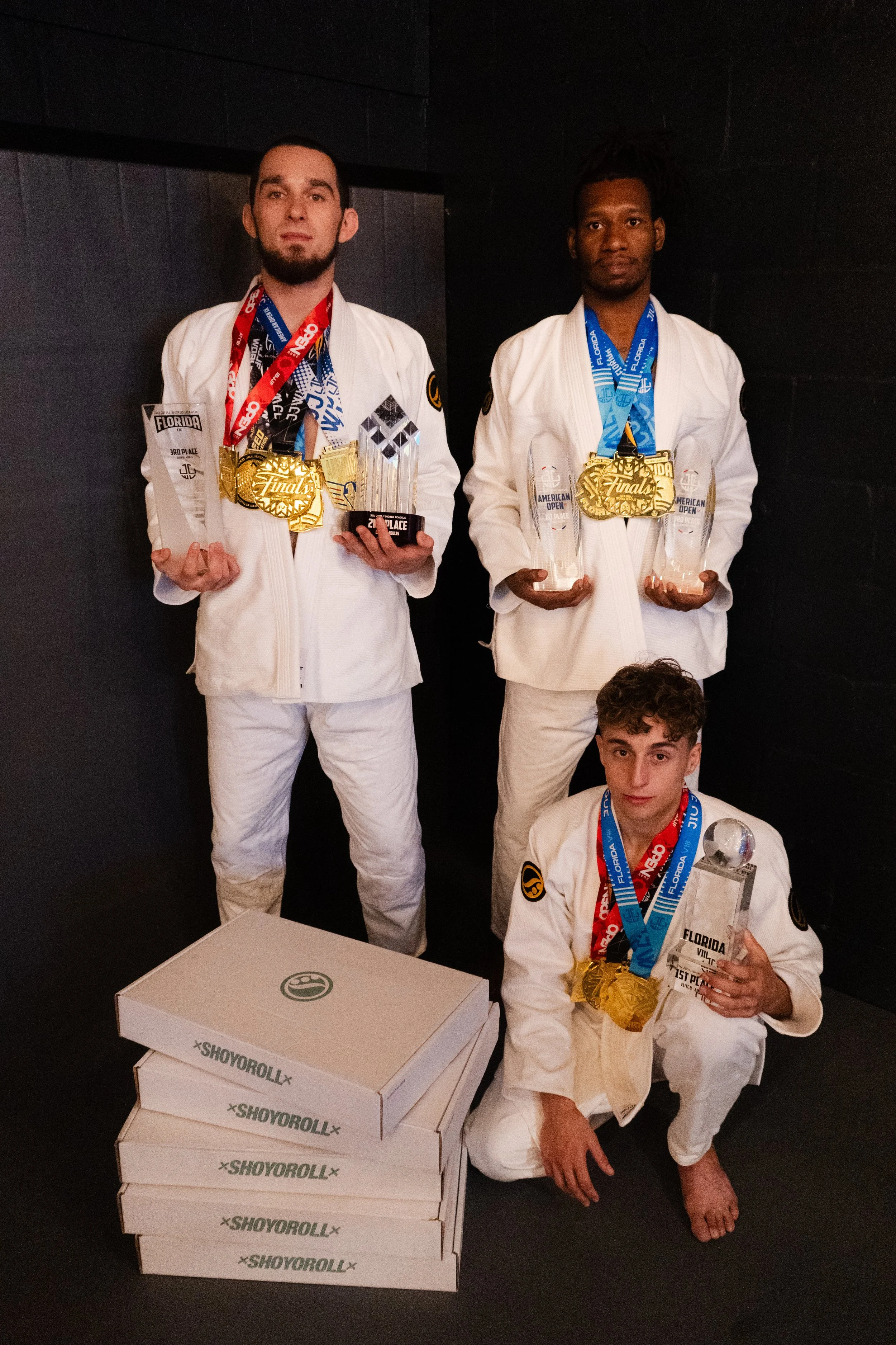 Three young men in white jiu-jitsu gis with medals and trophies pose after a competition. The two standing men hold glass and trophy awards, while the kneeling man holds a glass award. Three stacked boxes are in front of them.