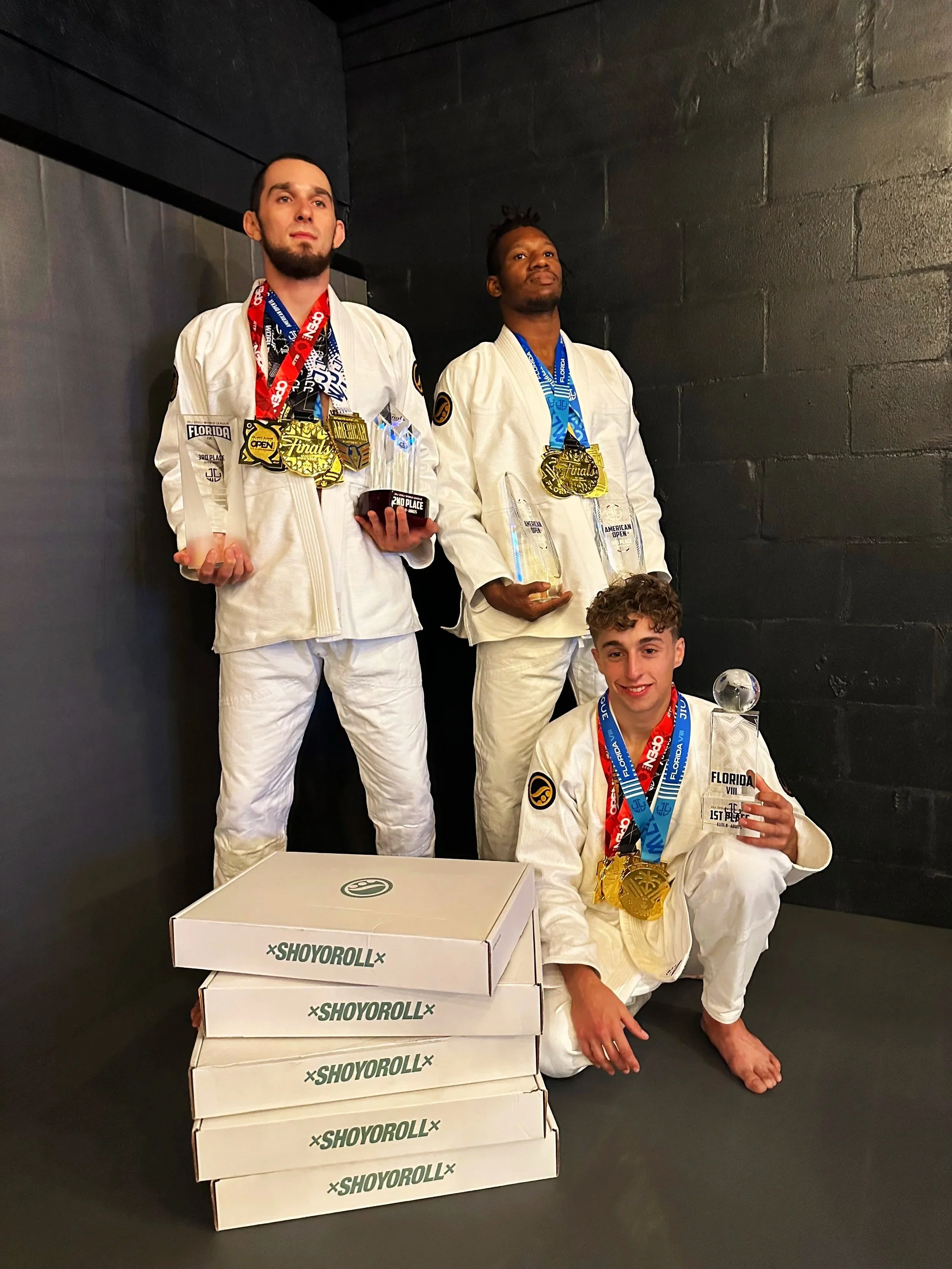 Three young men in white jiu-jitsu gis with medals and trophies pose after a competition. The two standing men hold glass and trophy awards, while the kneeling man holds a glass award. Three stacked boxes are in front of them.