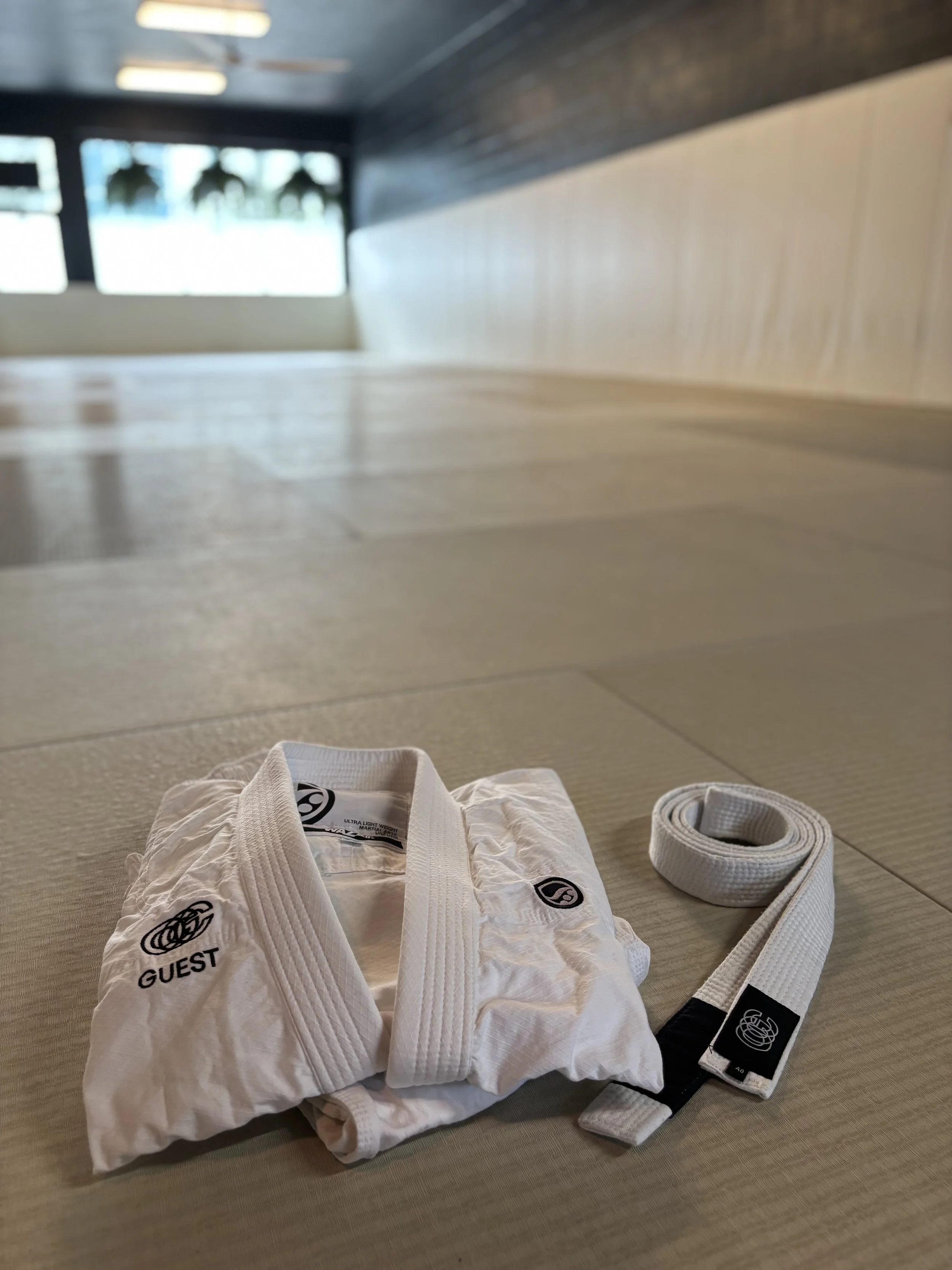 A white martial arts gi with a black and white belt, laid out on a light-colored mat in a gym or dojo with windows and natural lighting.