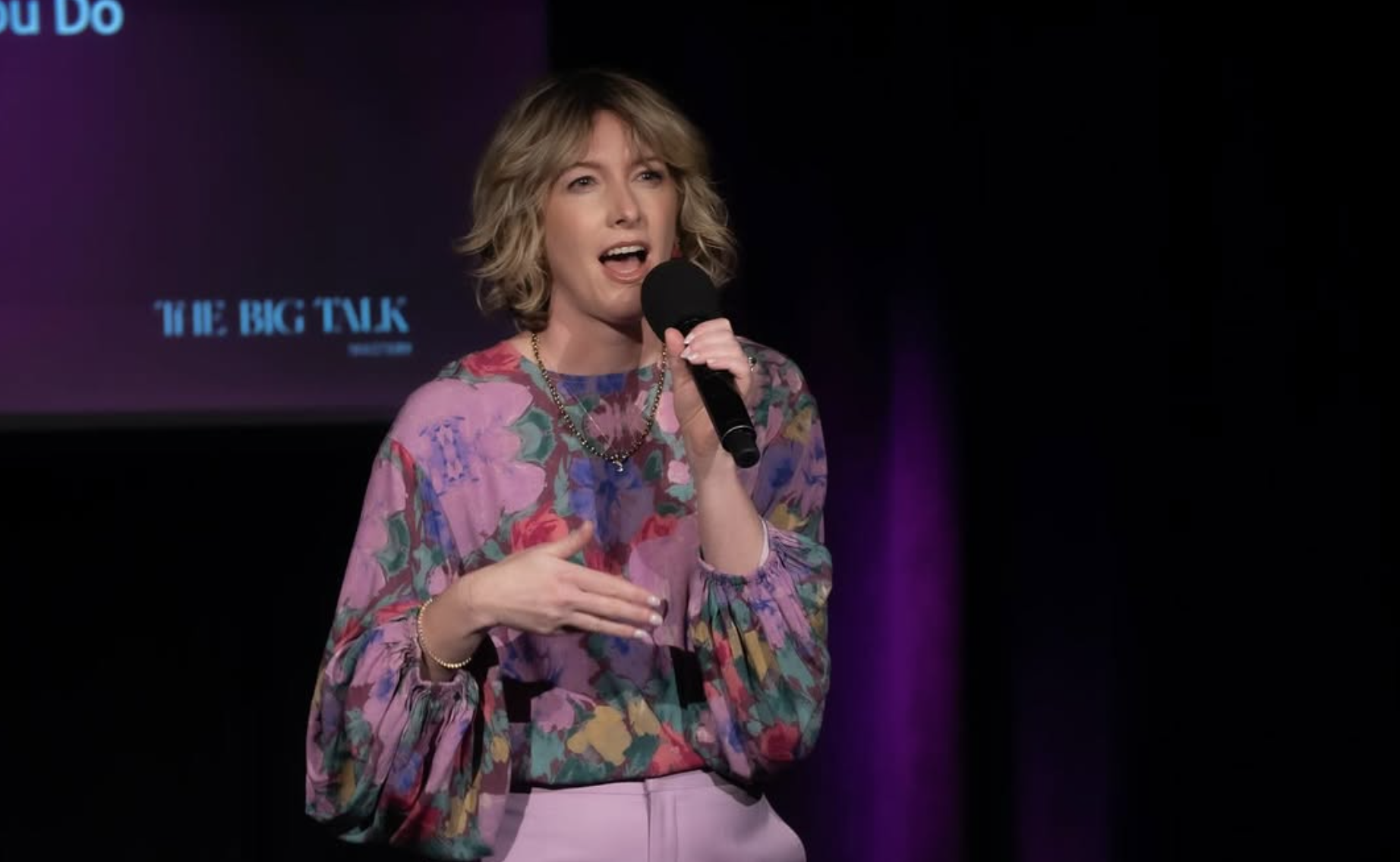 Natalie speaks into a microphone in a pink and floral outfit