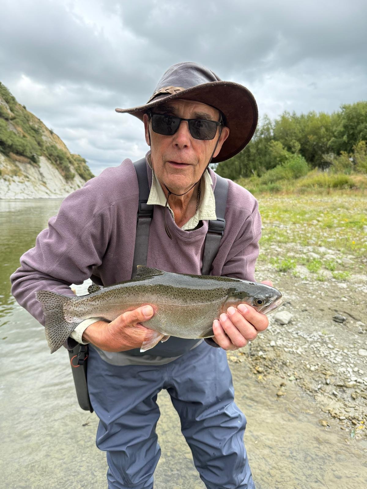 Fly fishing in New Zealand for brown and rainbow trout. Wellington, HawkesBay, Taihape, Central North Island. An older man wearing a wide-brimmed hat, sunglasses, and outdoor clothing, holding a fish.
