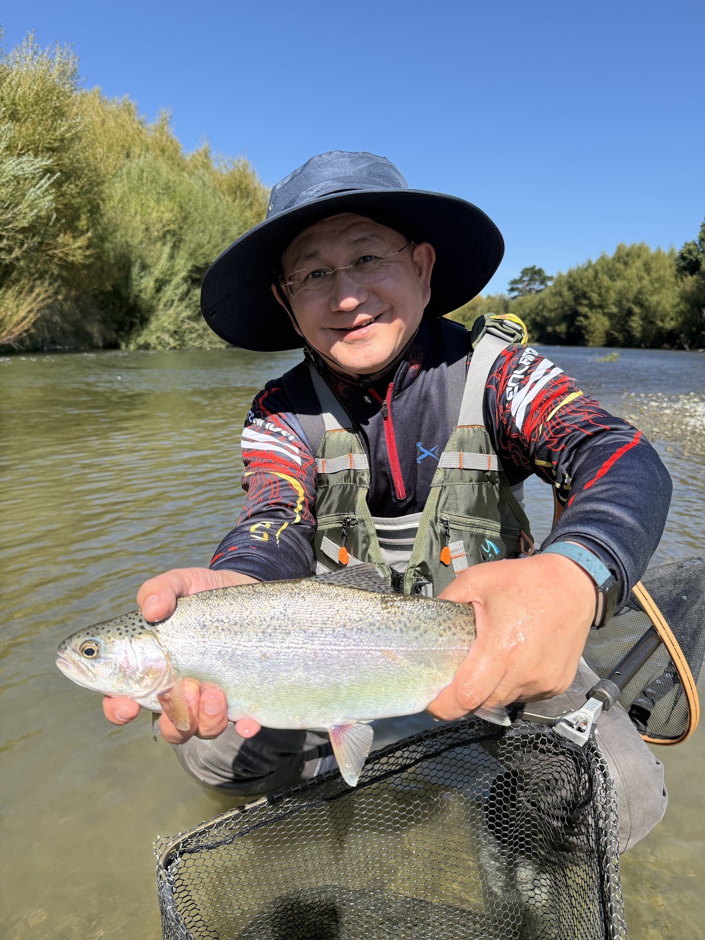 A fly fisherman in fishing clothing and a wide-brimmed hat holding a nice rainbow trout in a shallow part of the river with trees and blue sky in background. Euro Nymphing for trout in Pristine rivers in the North Island of New Zealand
