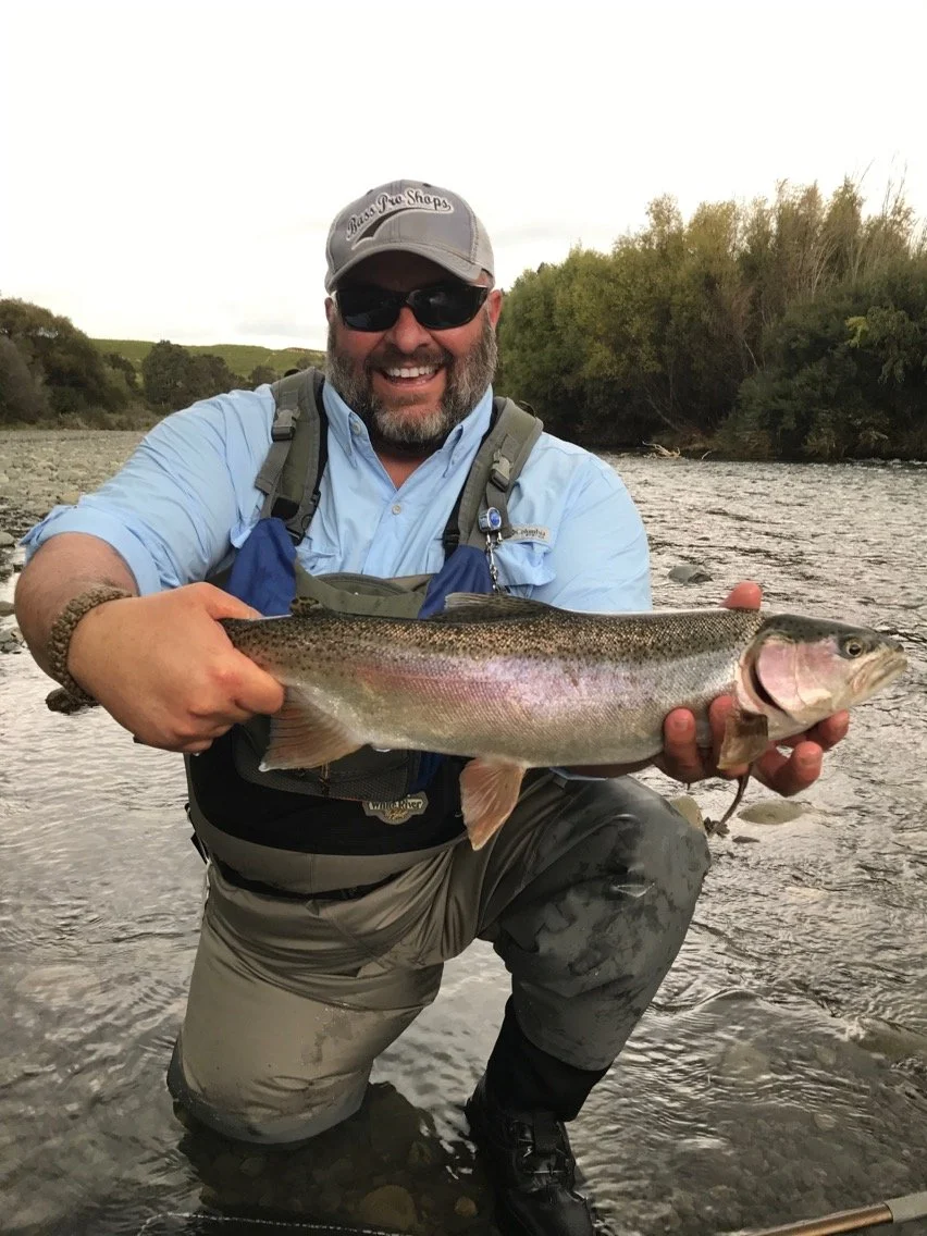 A fly fisherman holding a large rainbow trout fish in a river with trees in the background. Ultimate fishing experience with large rainbow and brown trout in HawkesBay, New Zealand