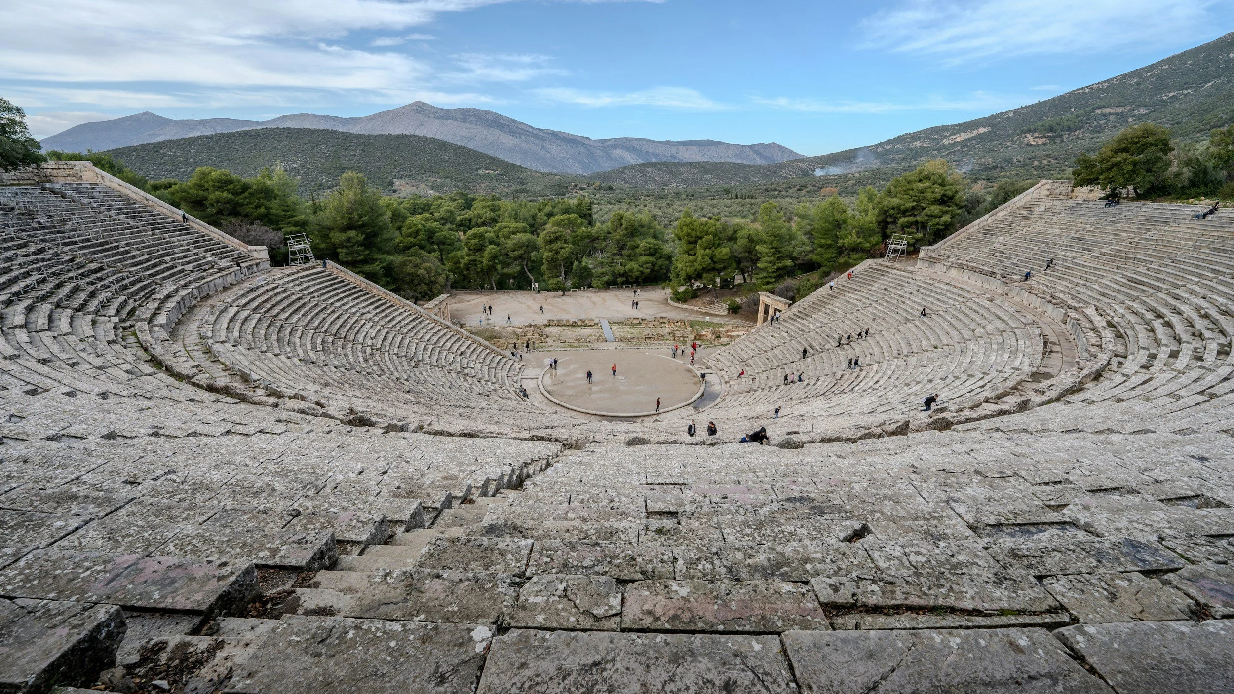 Panoramic view of the ancient theater of Epidaurus in Greece, with stone seating curving around the stage and mountains in the background — a UNESCO site dedicated to Asclepius, god of healing
