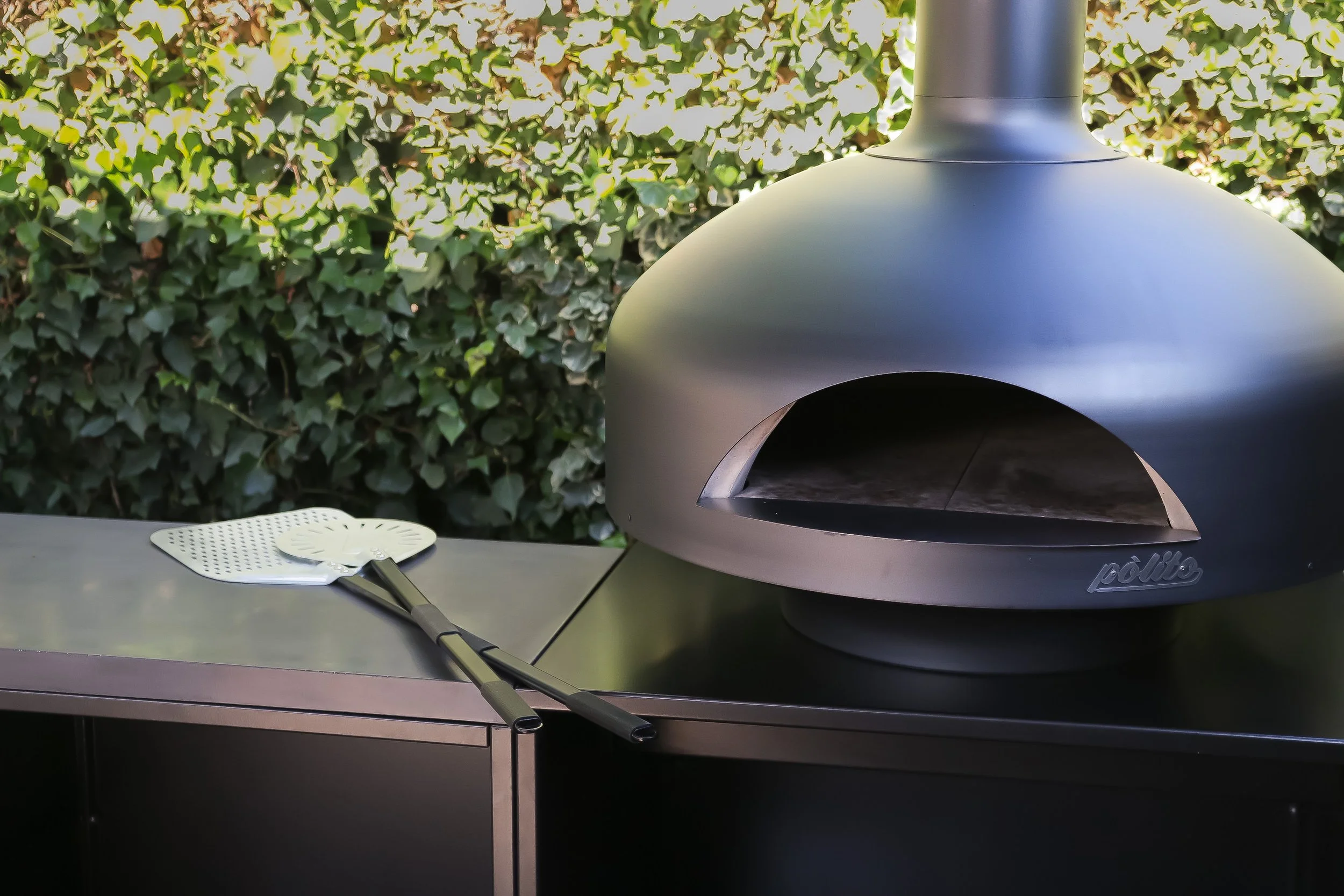 Outdoor pizza oven on a stainless steel table with pizza peels nearby, green foliage background.