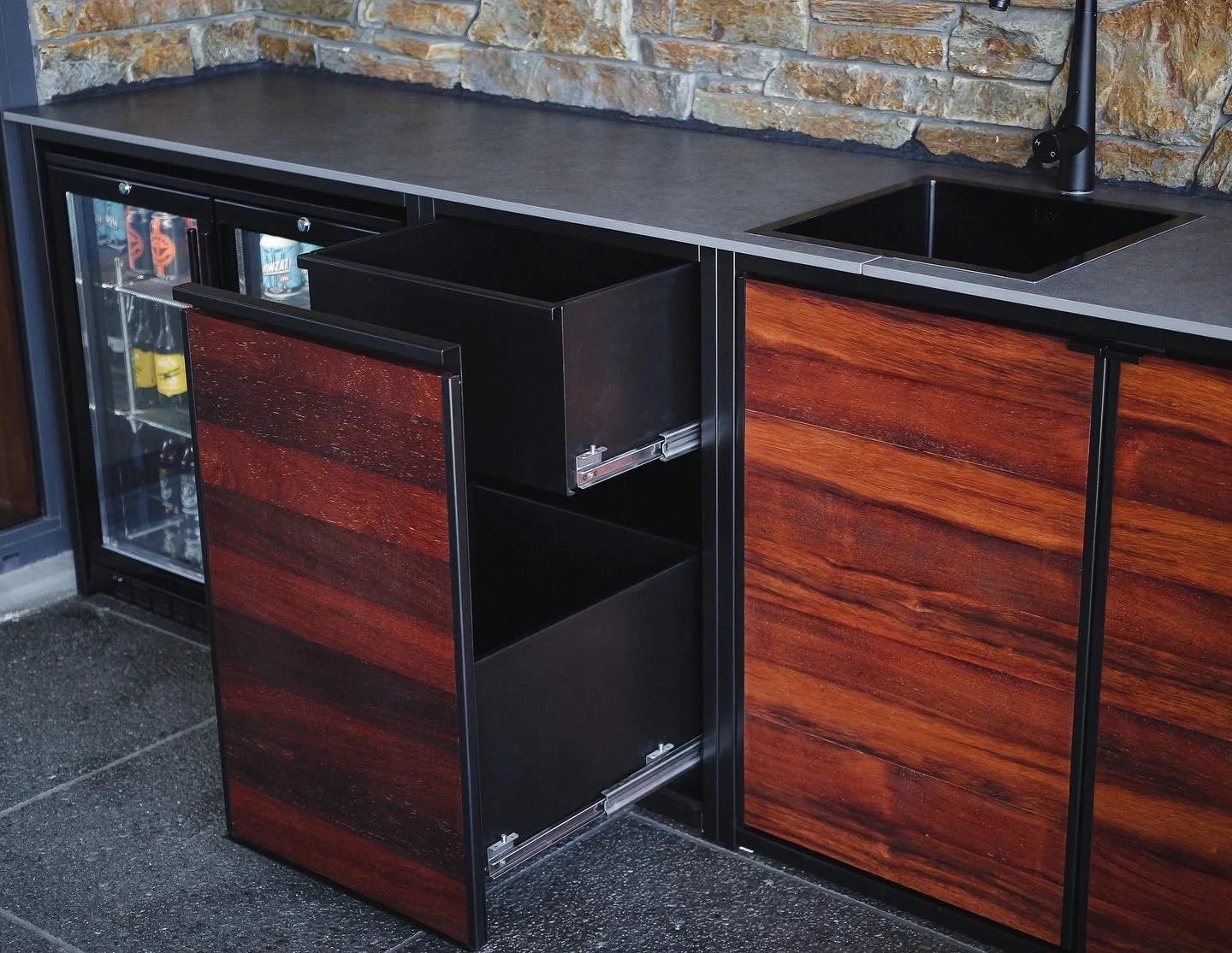 Bar area with dark wood cabinets, a stone wall, a gray countertop, a small fridge with beverages, black sink with faucet, and open cabinets.