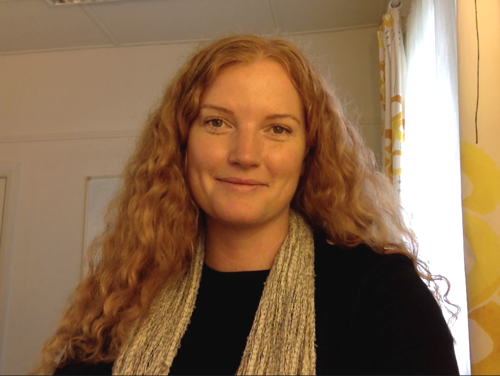 A woman with long, curly red hair smiling indoors near a window with a yellow and white curtain.