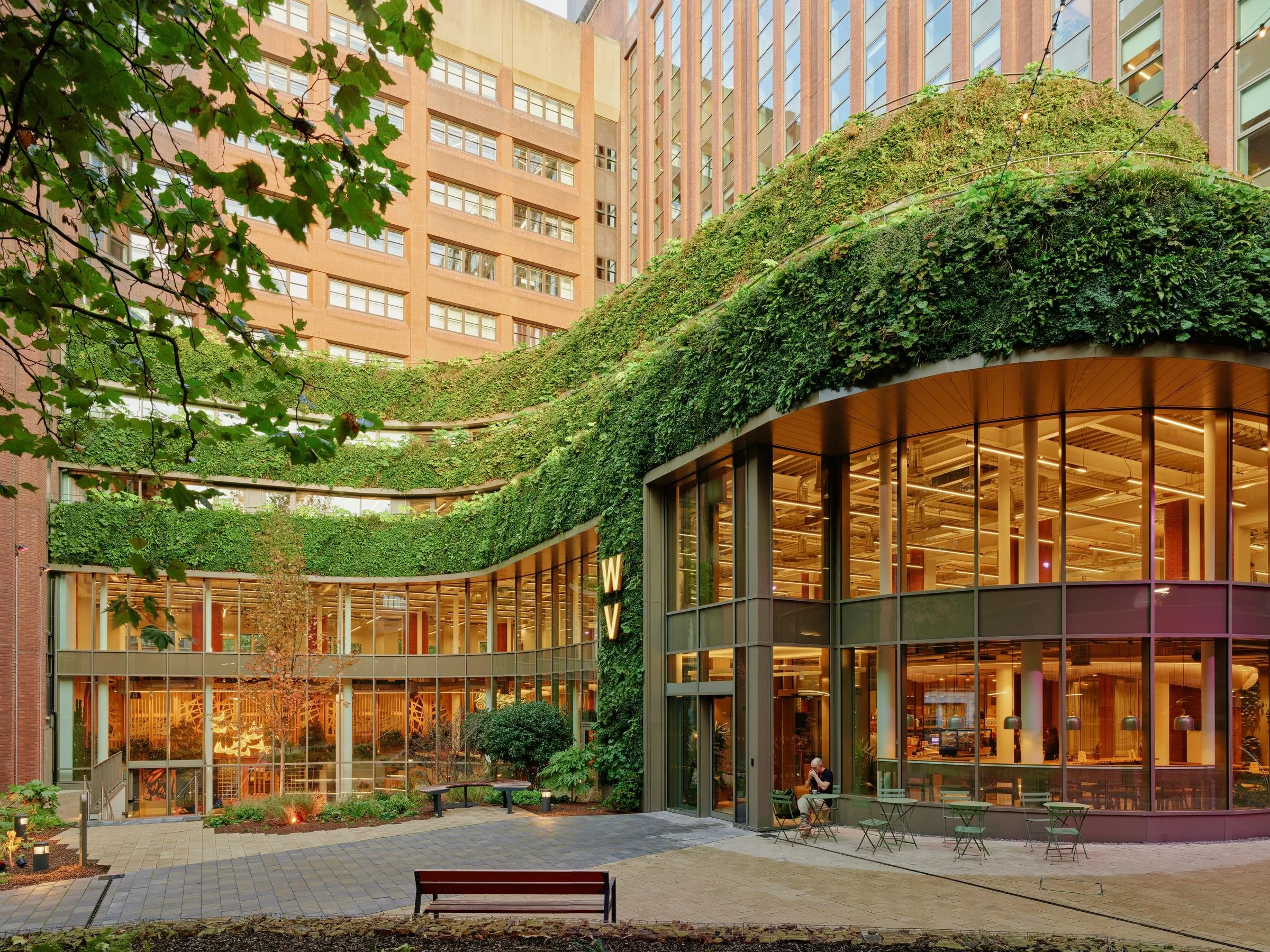 A modern building with large glass windows and a curved greenery-covered rooftop terrace. There is an outdoor seating area with tables and chairs in the foreground, and trees with green leaves surrounding the area.