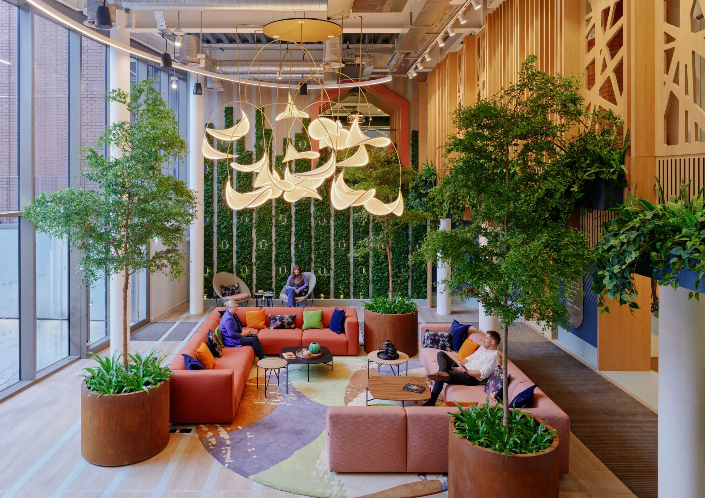 Modern indoor lounge area with large windows, green plants in large wooden planters, colorful sofas, and unique ceiling lighting.
