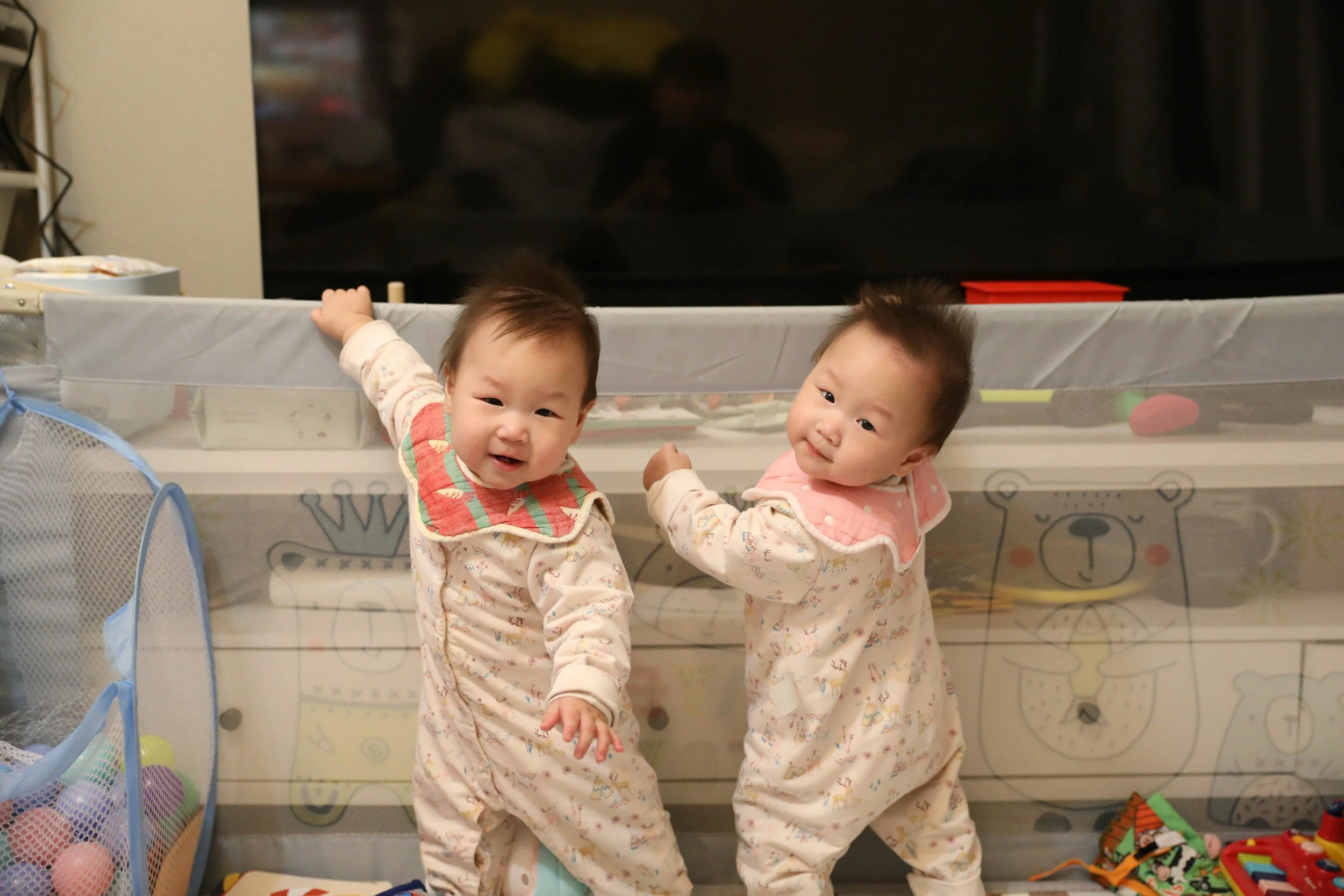 Two young children in pajamas playing near a protective playpen with toys, a TV, and a shelf in the background.