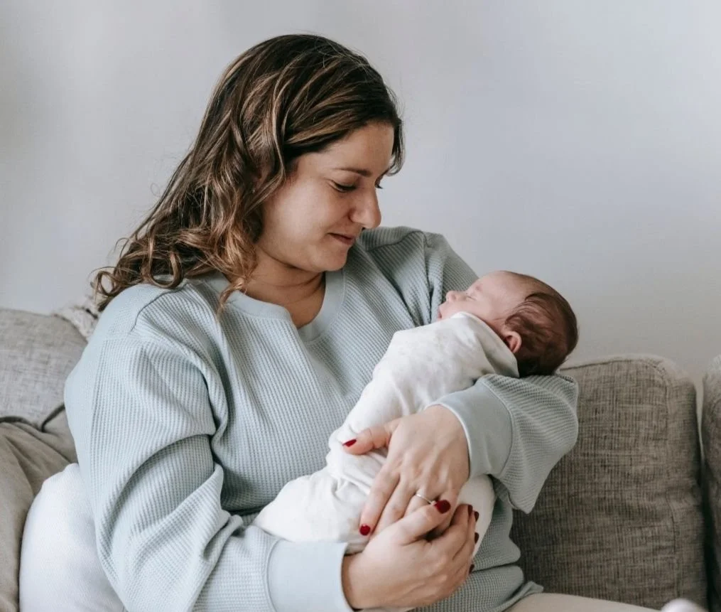 A woman holding a newborn baby indoors, sitting on a sofa.