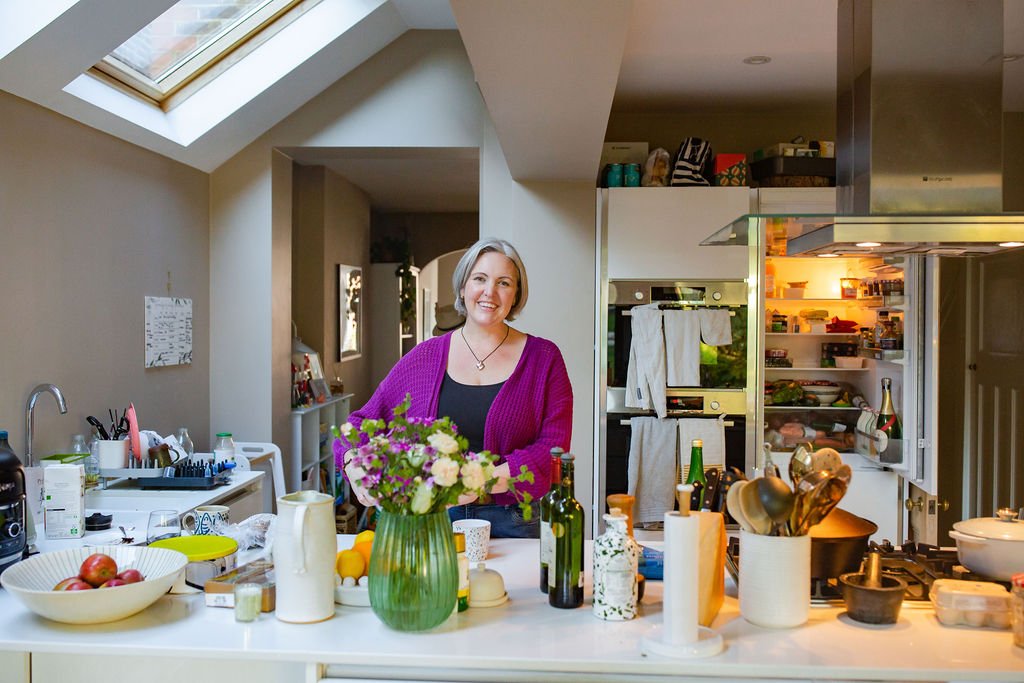 Sarah smiling in a kitchen with a white countertop filled with a vase of flowers, bottles, and various kitchen items. The background shows a refrigerator with open shelves and a skylight overhead.