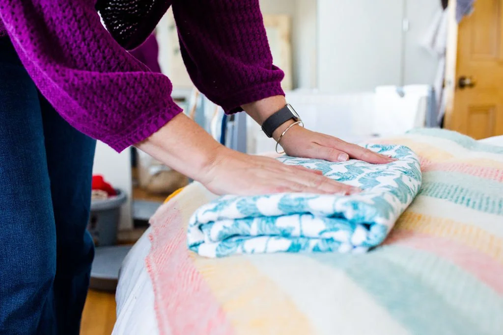 A doula to fold your laundry. Help for your postpartum days. Person folding a blanket on a bed in a bedroom.