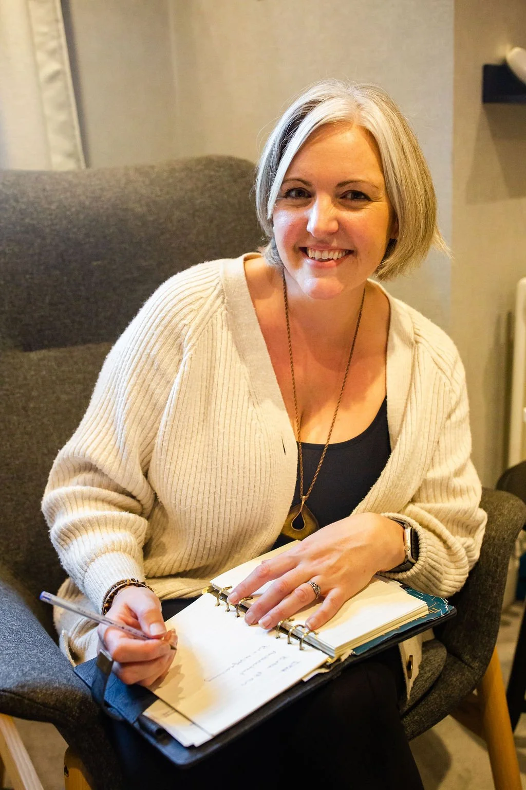A smiling woman with short blonde hair, wearing a beige cardigan and a black top, sitting on a dark gray armchair, holding a pen and a spiral notebook with papers.