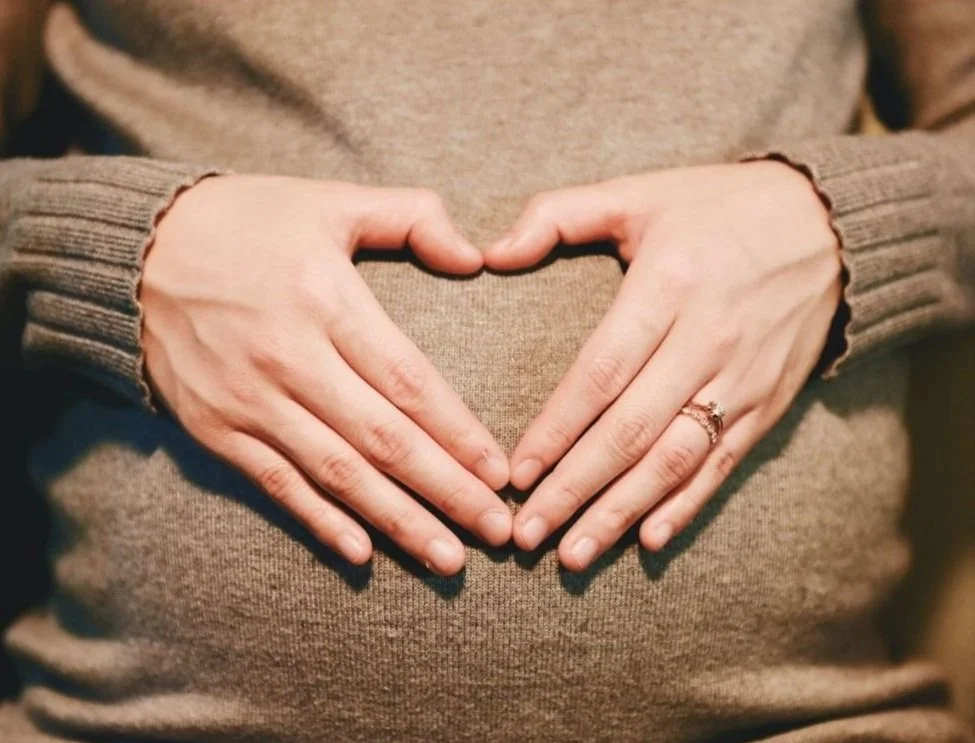 Two hands forming a heart shape on a pregnant belly, with a ring visible on the right hand.