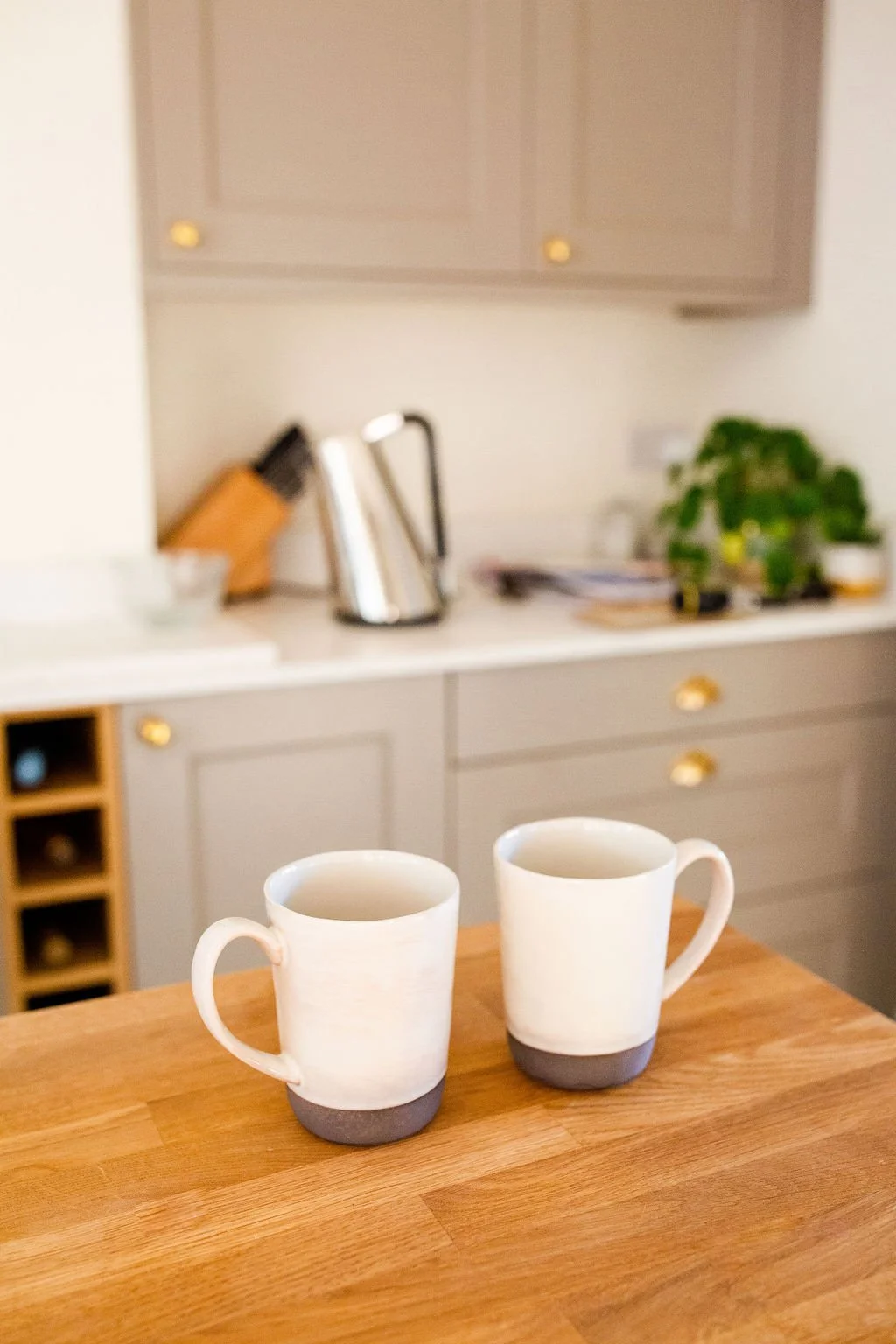 Someone to make you a cup of tea. Two white ceramic coffee mugs with grey bases on a wooden kitchen countertop.