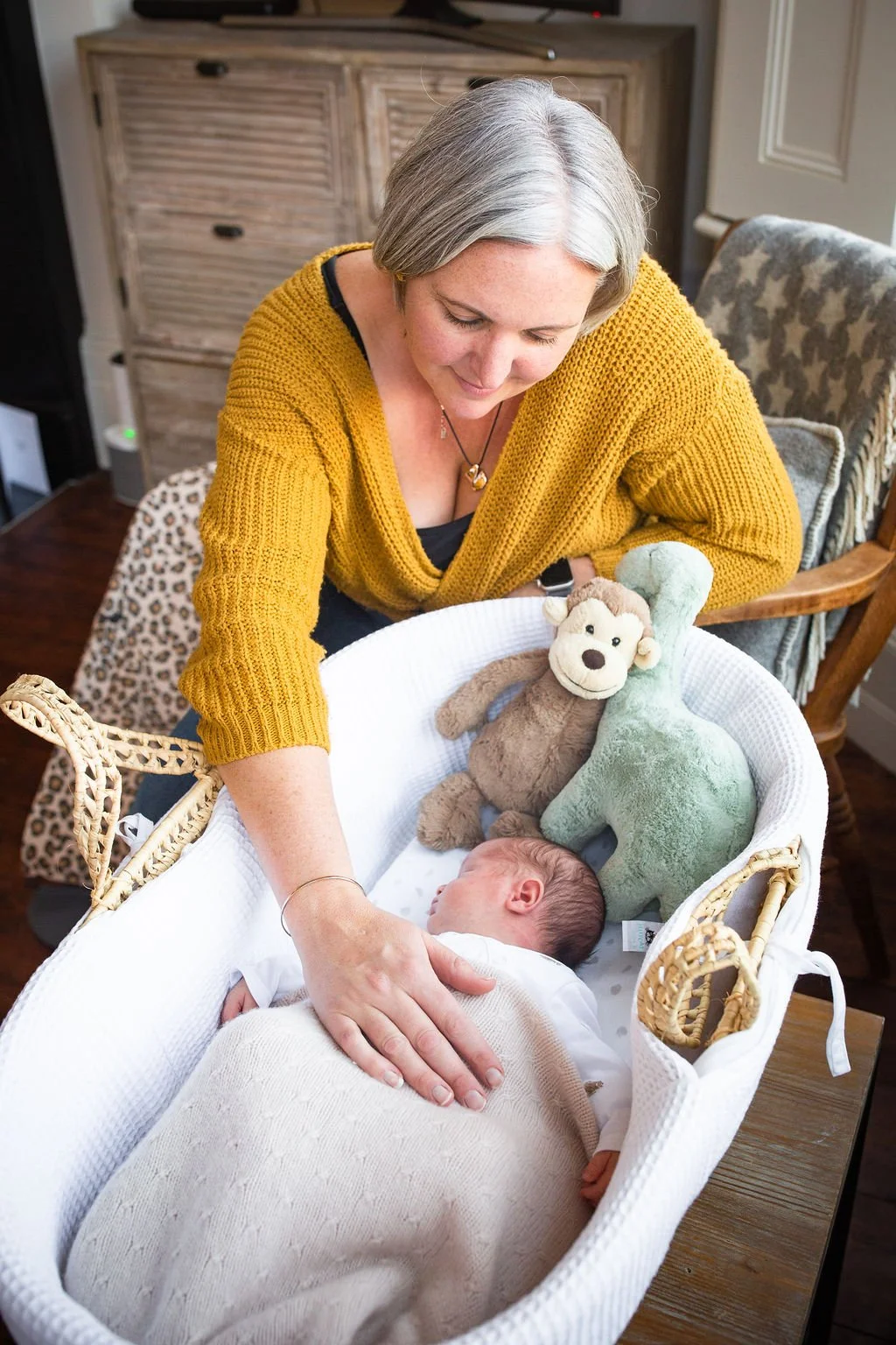 A postnatal doula and a mustard yellow cardigan smiling at a sleeping baby in a bassinet, surrounded by plush toys of a monkey and a dinosaur.