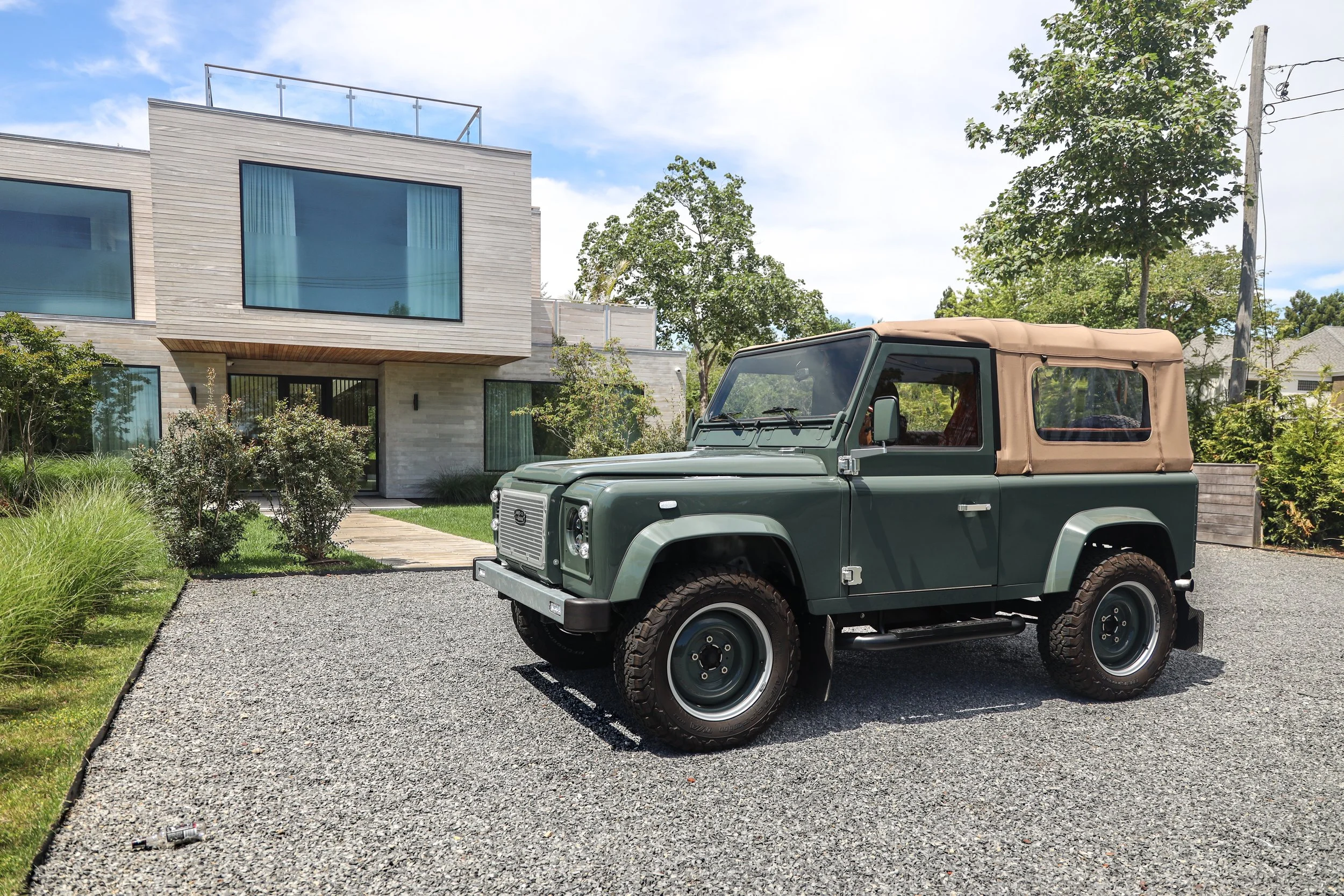 A classic green Land Rover Defender with a tan soft top parked on a gravel driveway in front of a modern house surrounded by greenery.