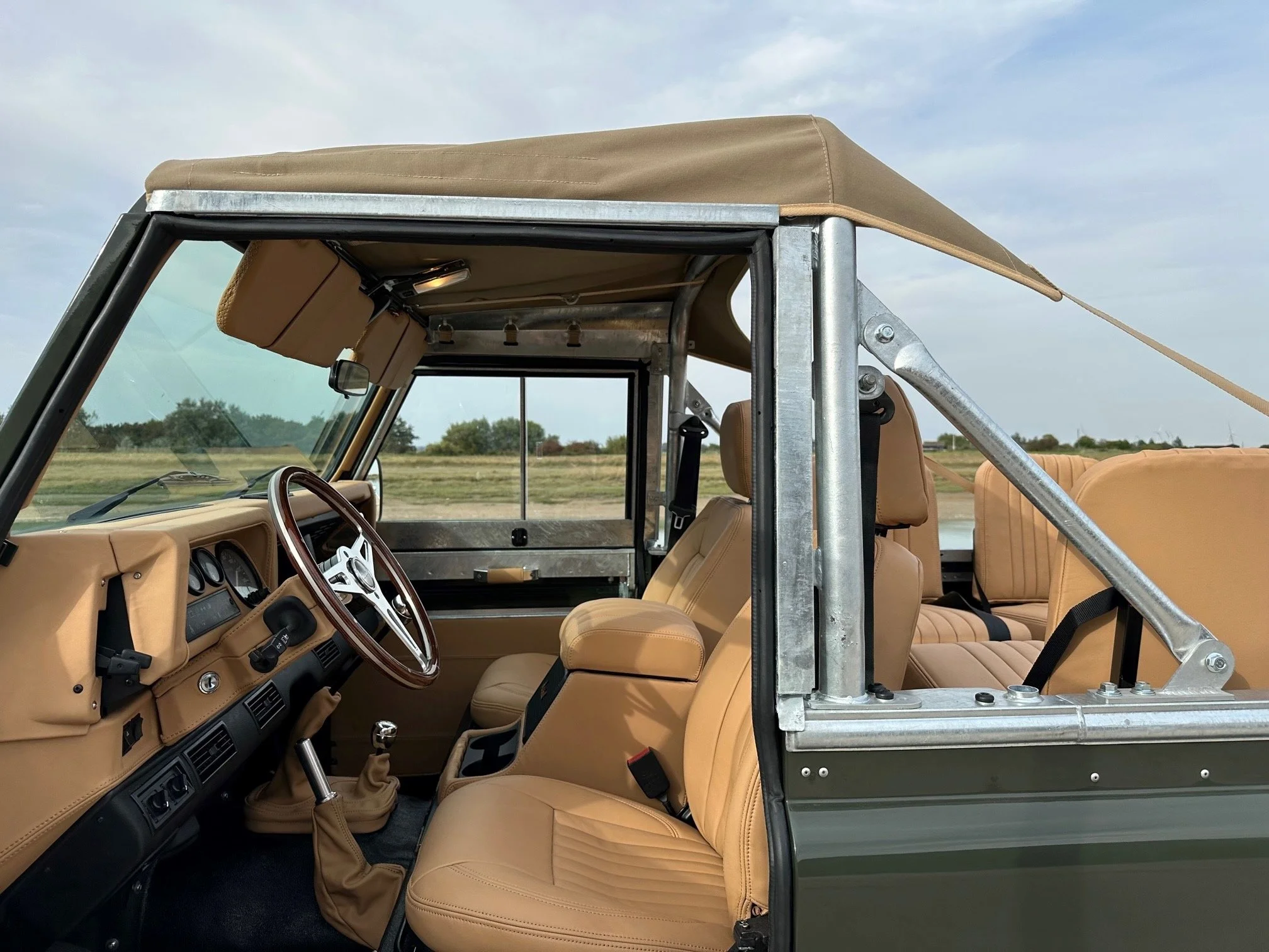 Interior of a vintage off-road vehicle with tan seats and dashboard, metal framework, and an open back with a shaded canopy.