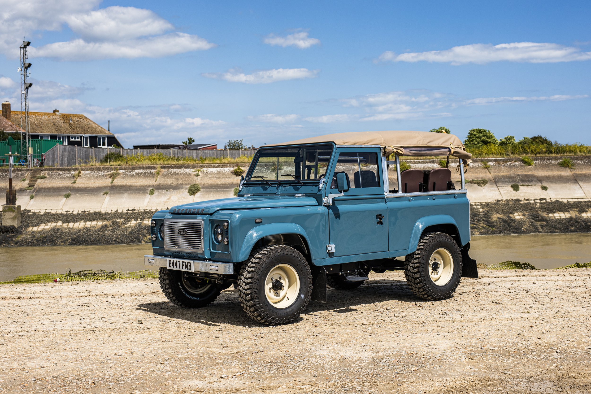 A vintage blue Land Rover Defender with beige soft top parked on a sandy surface near a river, under a partly cloudy sky.