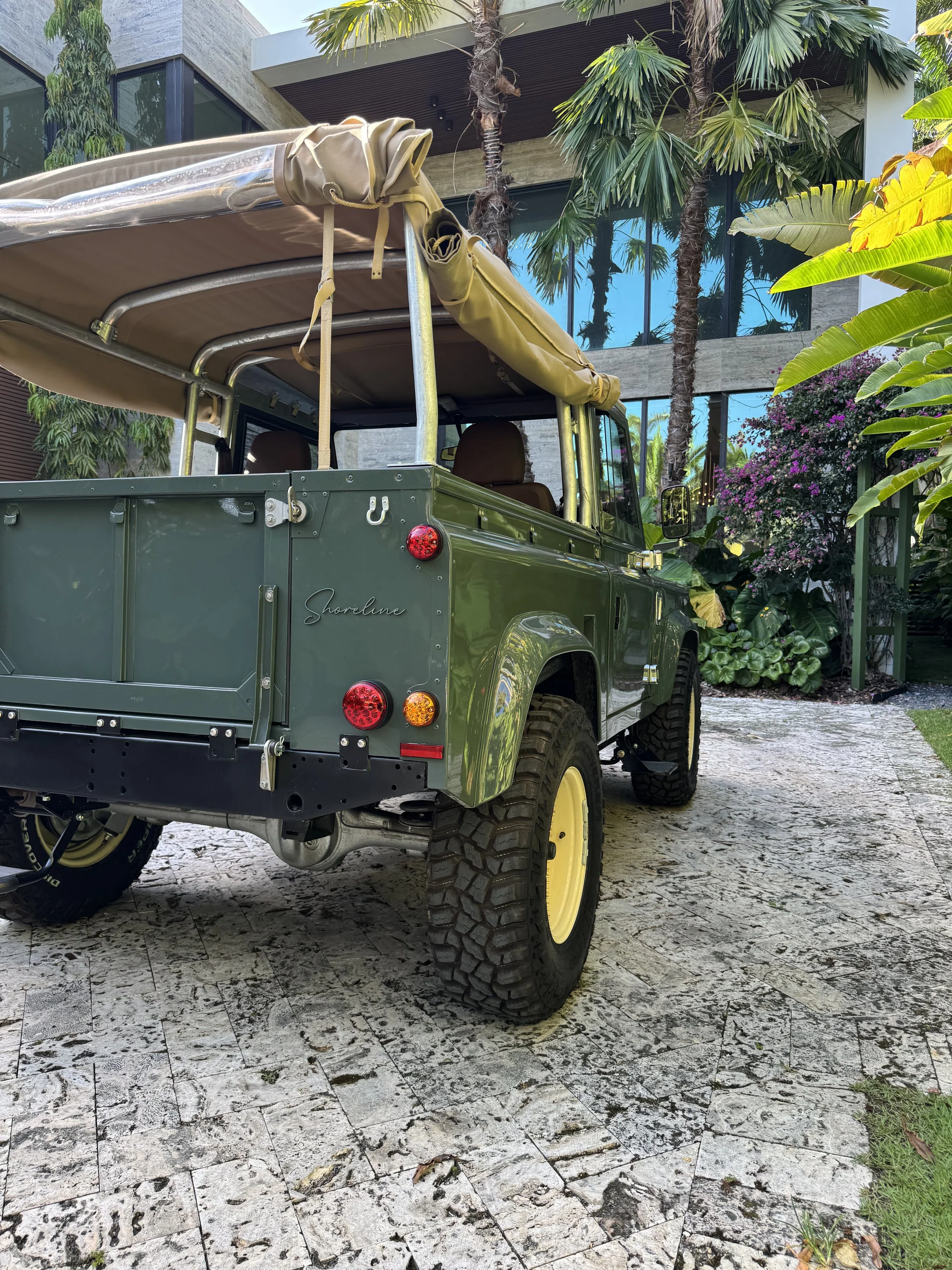 Vintage green Land Rover with yellow wheels parked on a stone driveway surrounded by tropical plants in front of a modern building with large glass windows.
