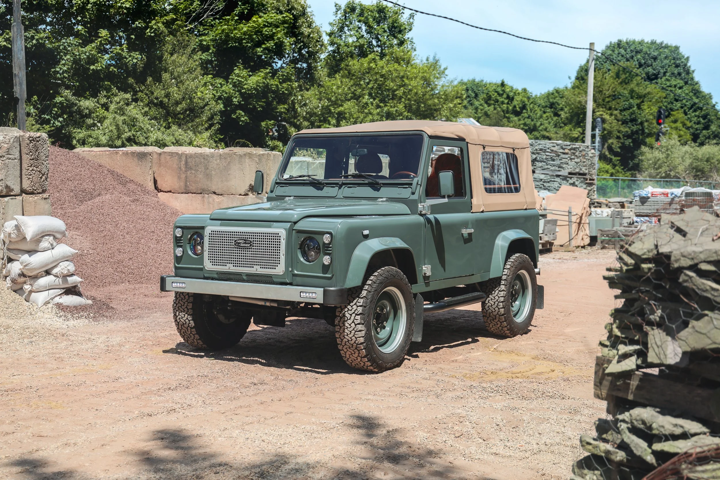 A vintage green Land Rover Defender with a tan soft top parked outdoors on dirt, surrounded by stacks of rocks, sandbags, and construction materials, with trees and utility poles in the background.