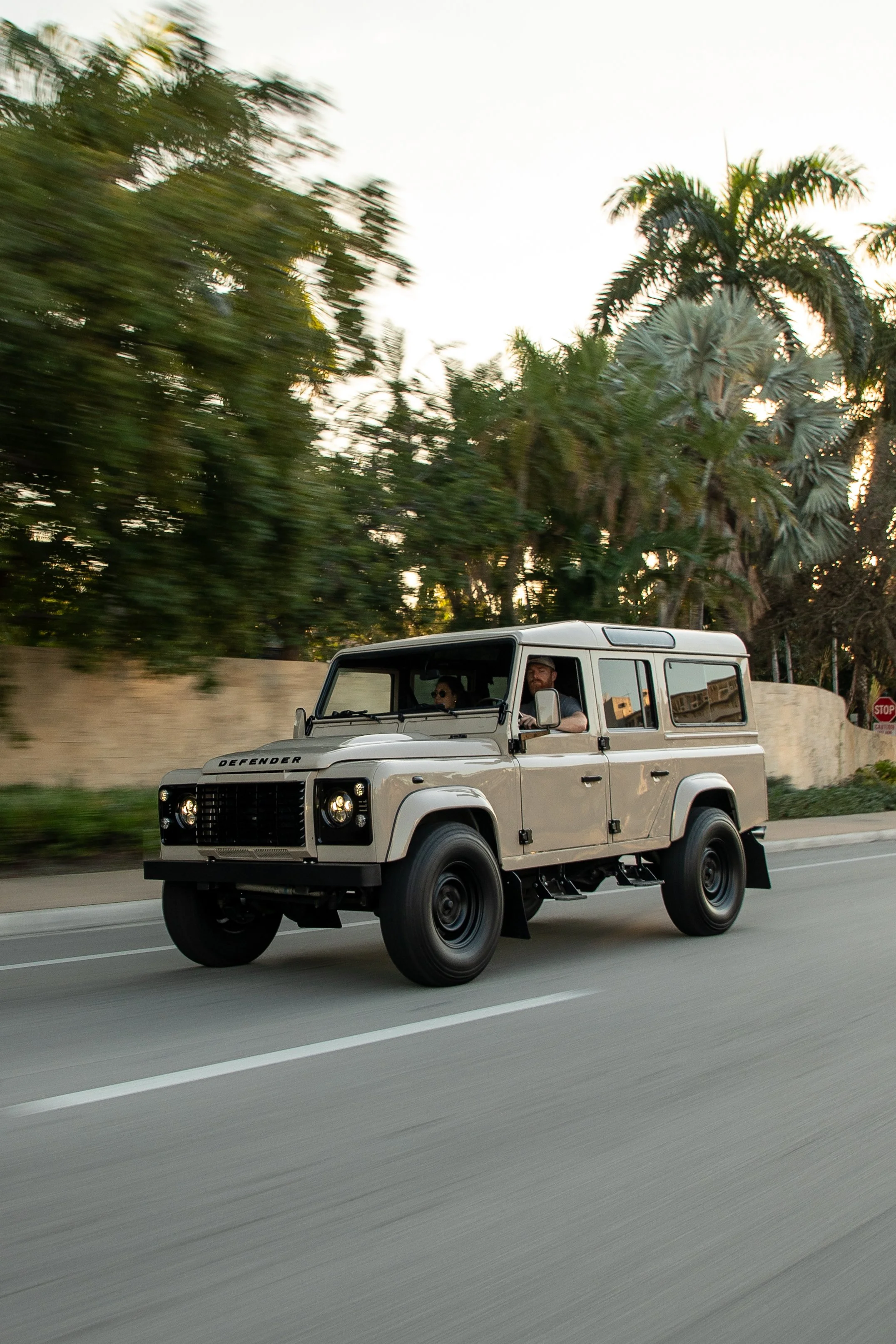 A beige Land Rover Defender SUV driving on a city street with palm trees in the background.