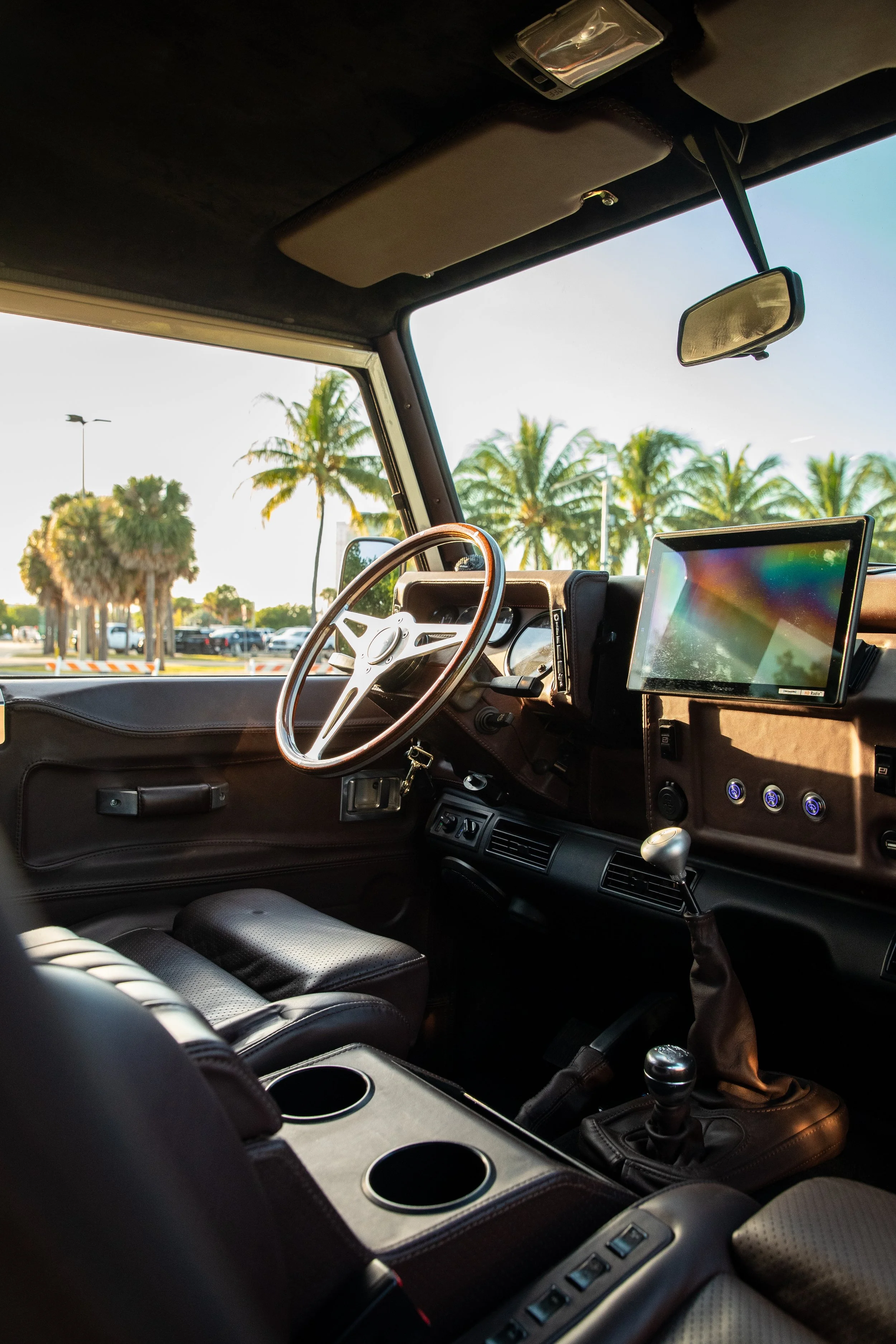 Interior view of a vehicle with a steering wheel, gear shift, dashboard, and a mounted display screen, with palm trees visible outside through the windshield.