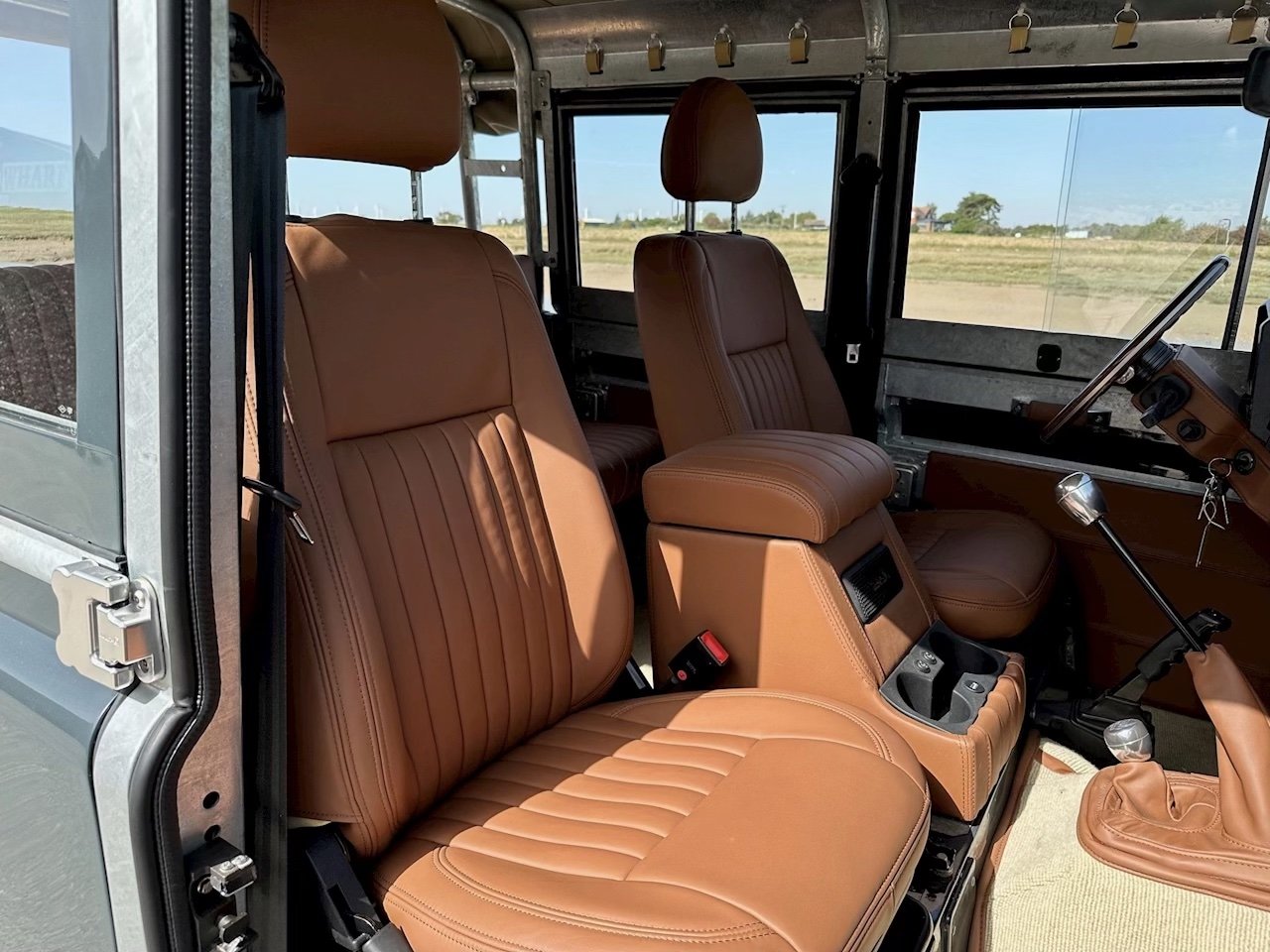 Interior of a vehicle with tan leather seats, gear shift, and dashboard, with an open window showing a rural landscape outside.