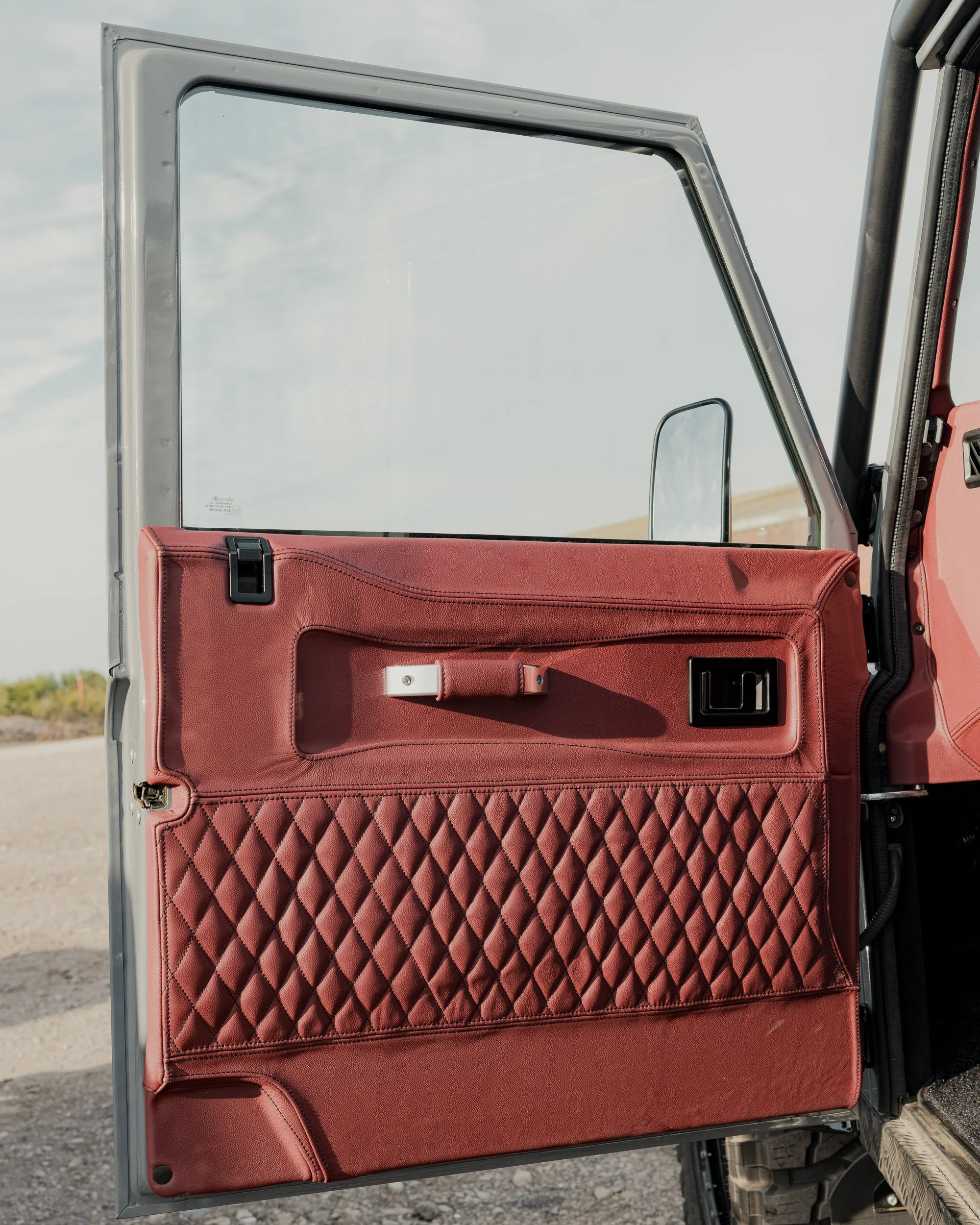 Close-up of a vintage car door interior with a red quilted leather panel, black handle, and window in a parked vehicle.