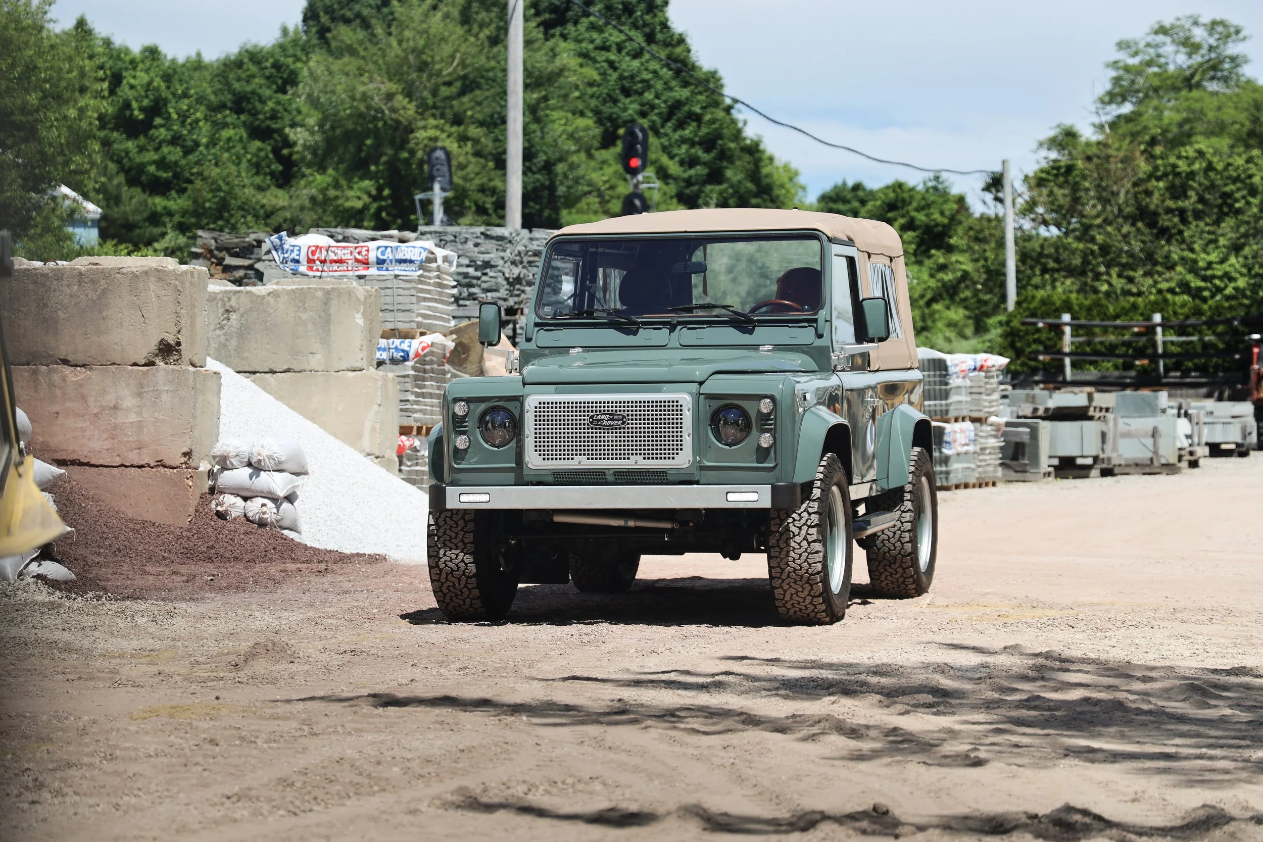 A green vintage Land Rover parked on a gravel surface in a construction or supply yard, with stacks of concrete blocks, gravel, and other building materials in the background, and trees and power lines beyond.