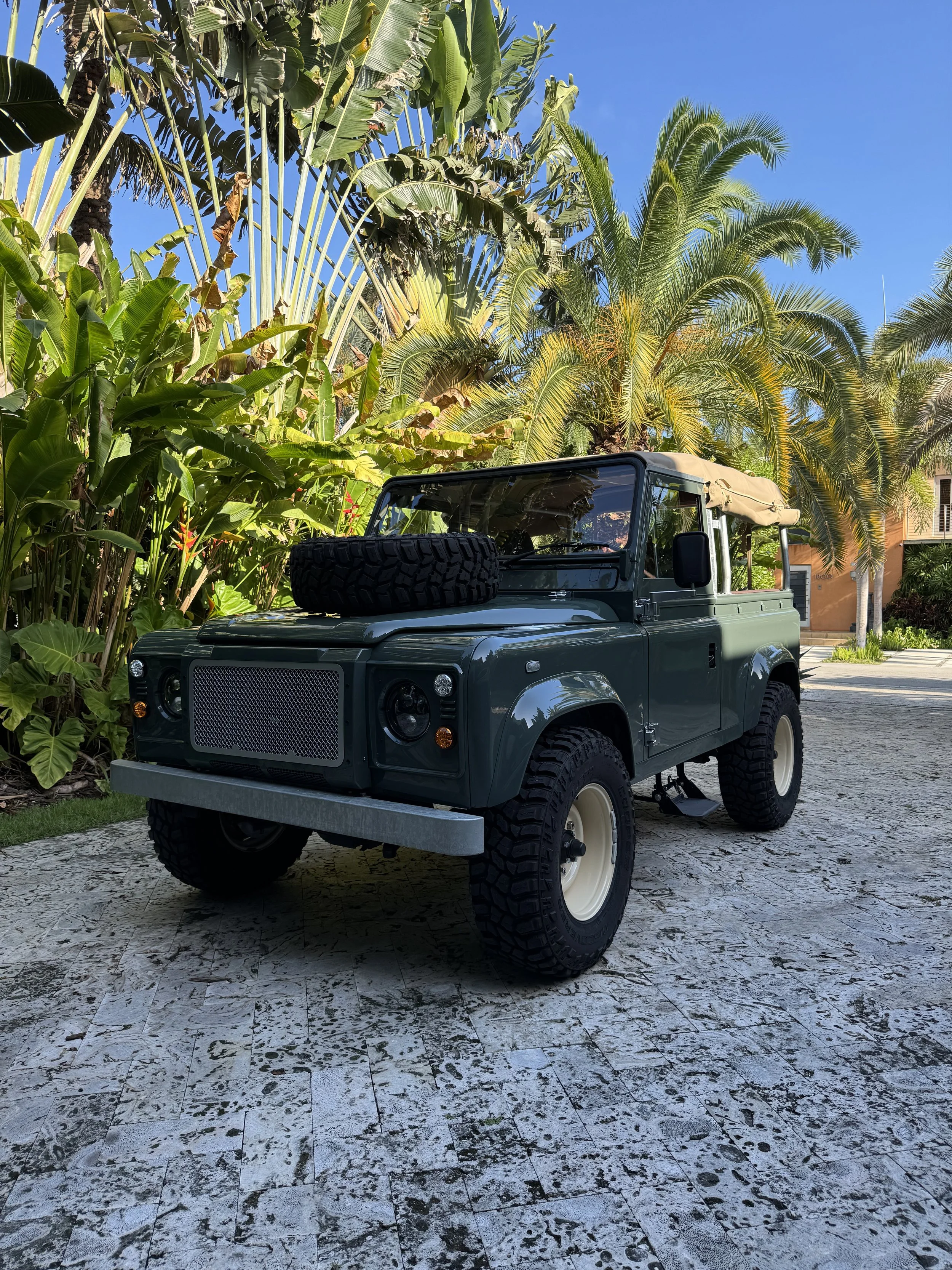 A vintage green off-road vehicle with a spare tire on its hood, parked on a stone driveway surrounded by lush tropical plants and trees, including palm trees, under a clear blue sky.